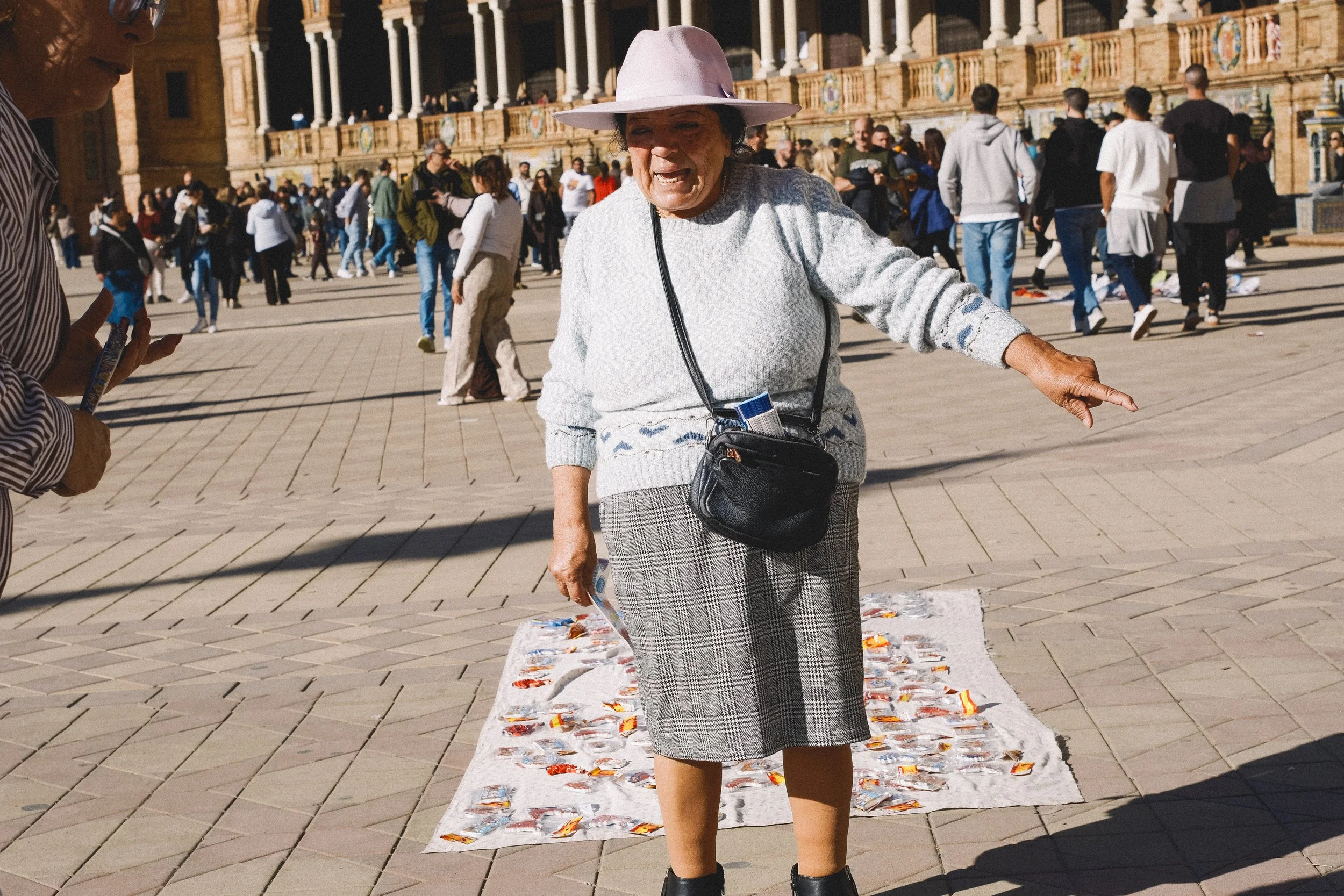 An elderly woman wearing a large hat, gray sweater, and plaid skirt stands on a large square in front of a historic building. She is smiling and pointing at a blanket with small packaged goods on the ground. There are many people in the background.