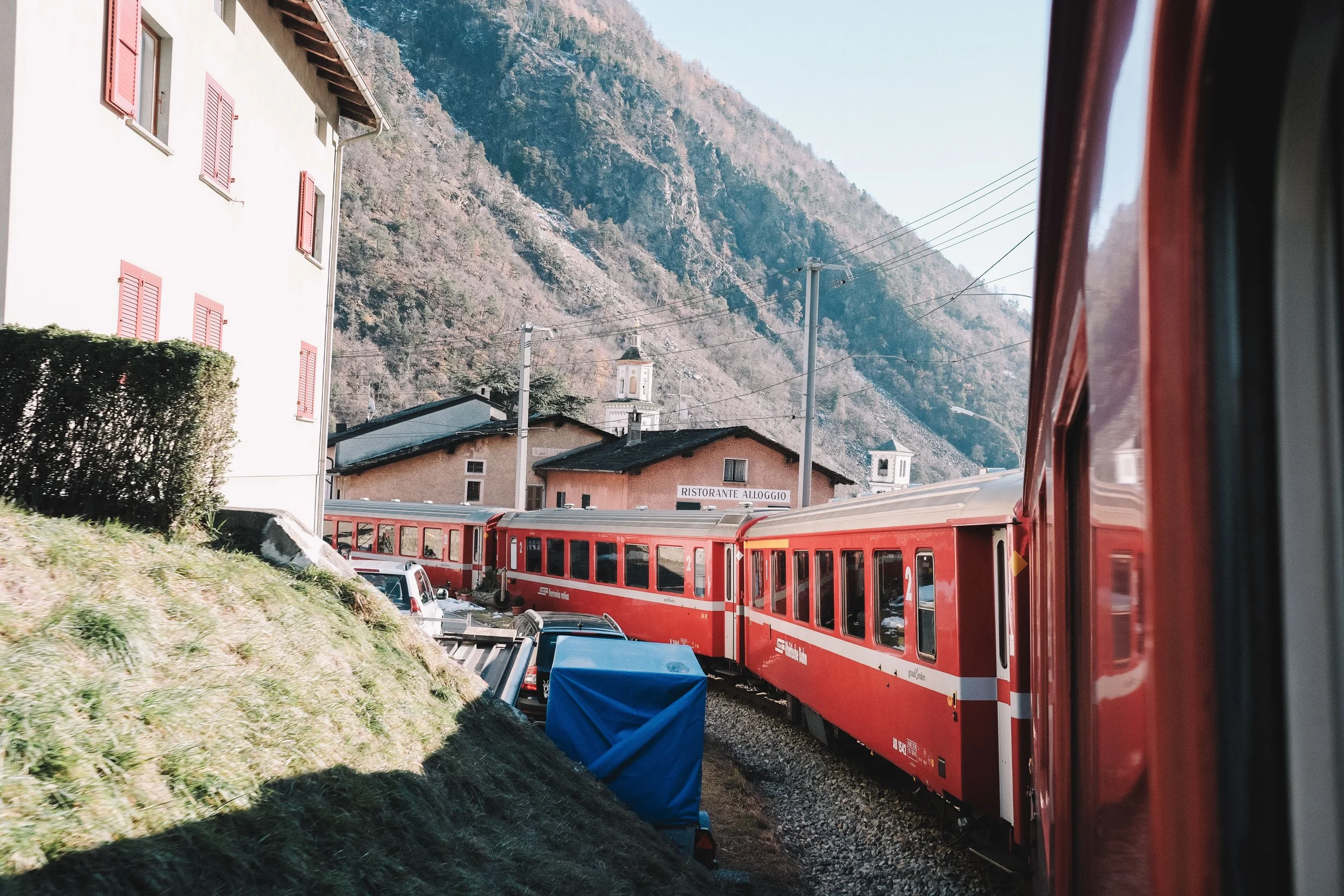Red train passing through a small town with a restaurant, surrounded by mountains and houses with red shutters.
