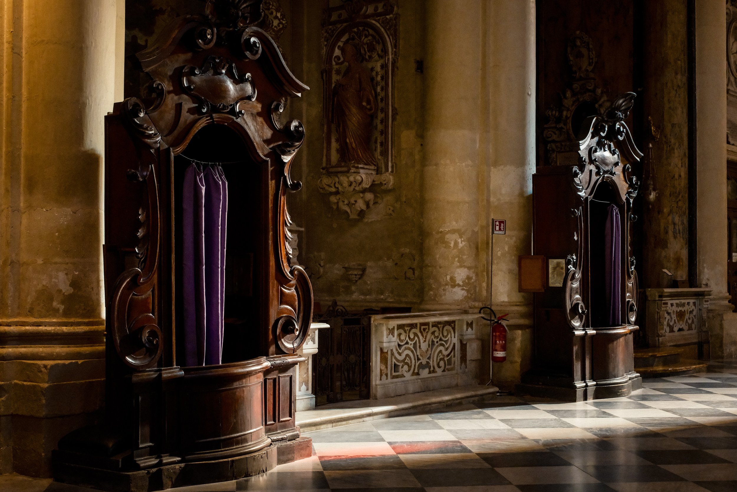 Interior of a historic building with two ornate wooden confessional booths draped with purple curtains, and a painted religious icon on the wall. The scene is dimly lit with stone columns and decorative elements.