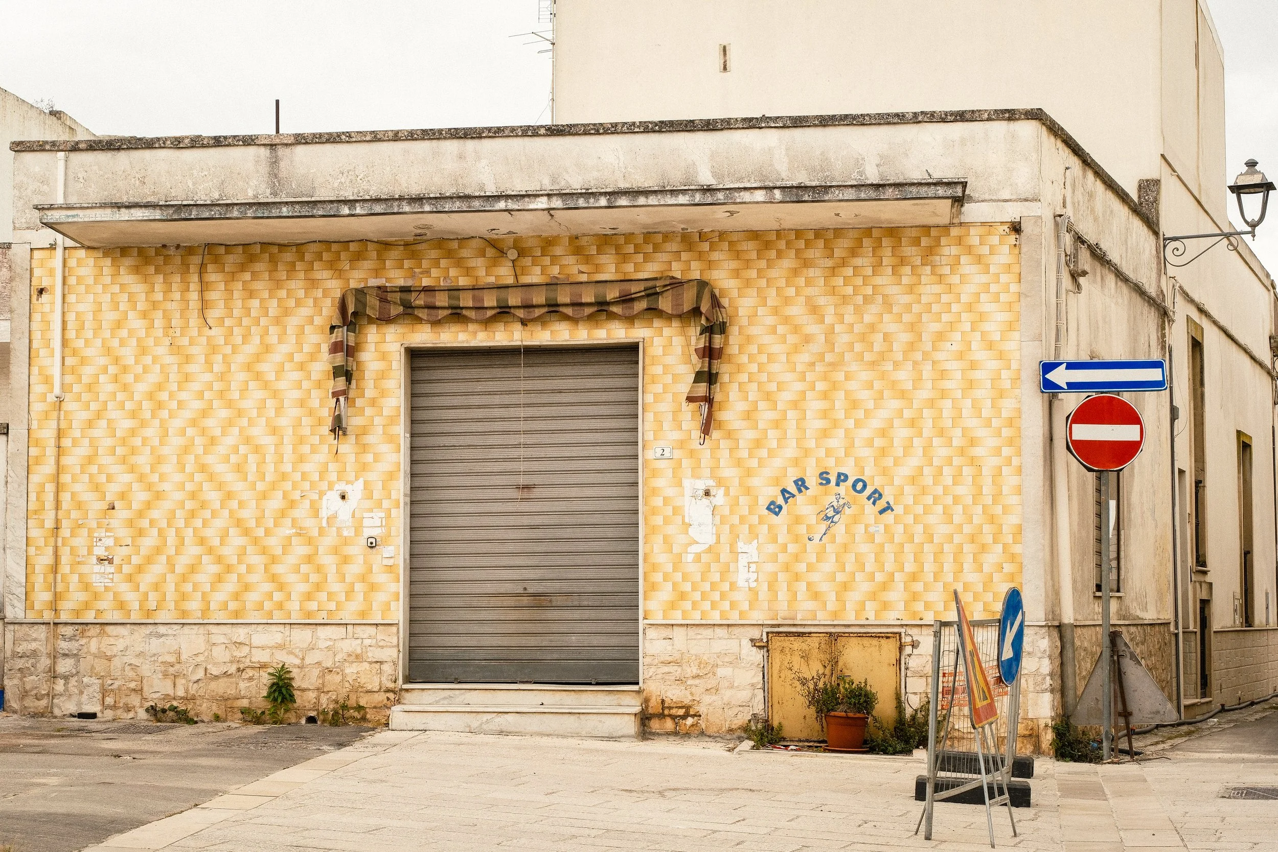 The exterior of a building with a yellow brick pattern, featuring a closed metal roll-up door, a patched wall, and some street signs nearby.