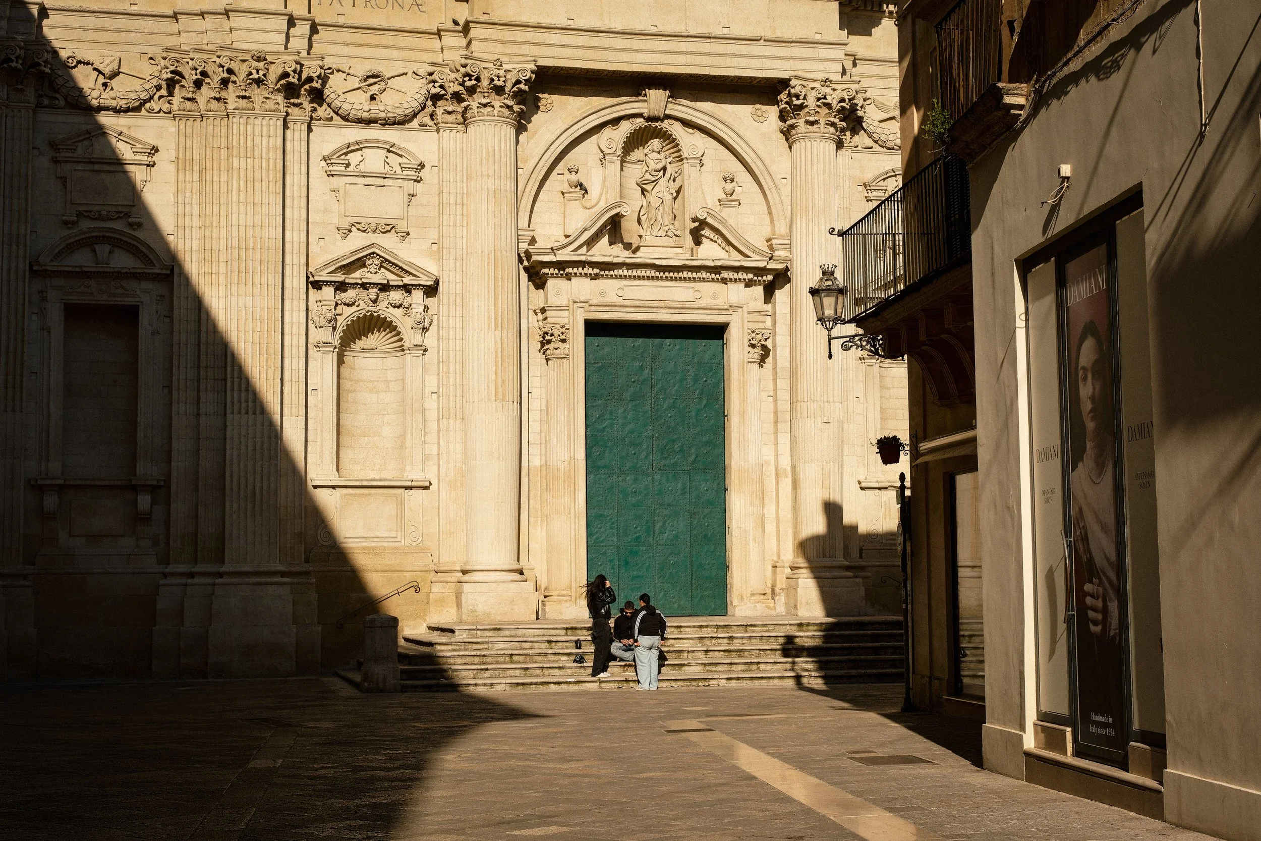 A historic stone building with ornate architectural details, including columns and sculptures, across steps where a few people are standing and sitting, with shadows cast on the building.