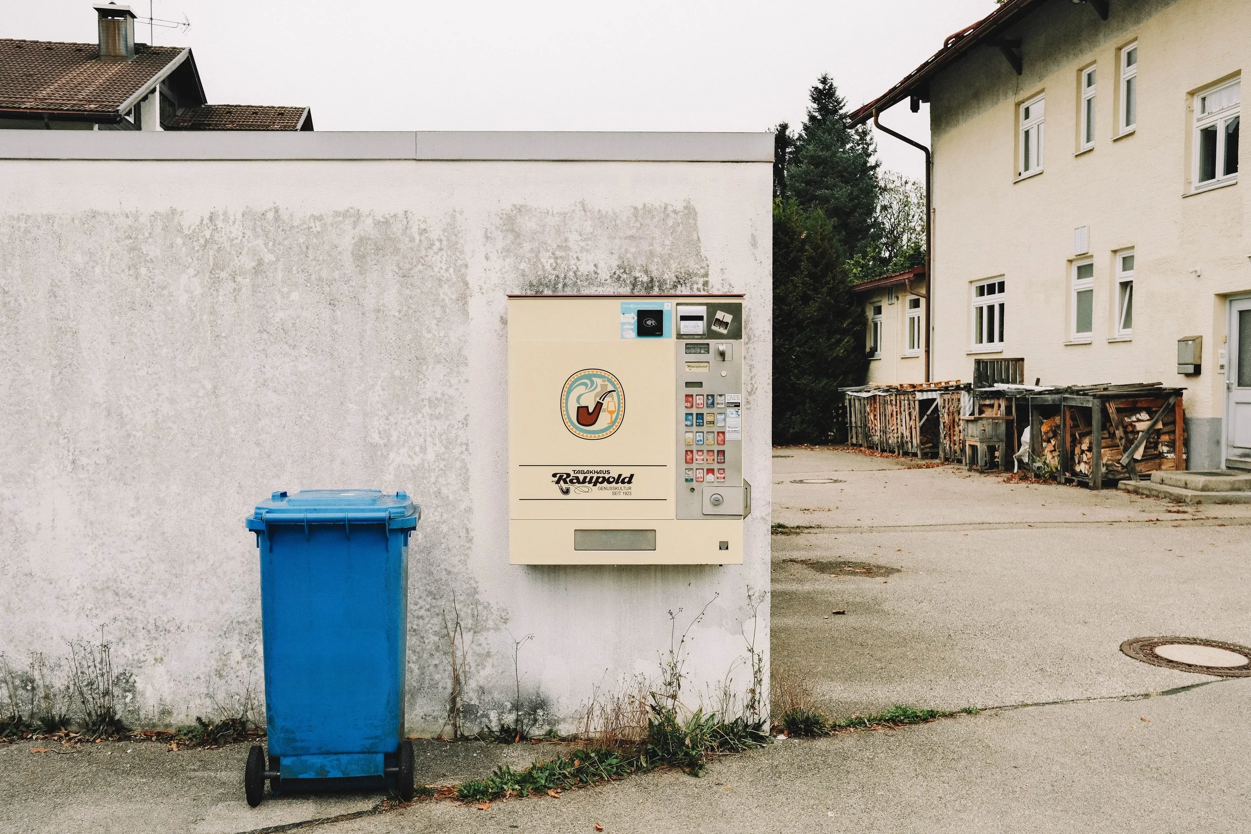 A cigarette vending machine mounted on a white wall next to a blue garbage bin on a paved area. To the right, there's a building with stacked wood and a small yard.