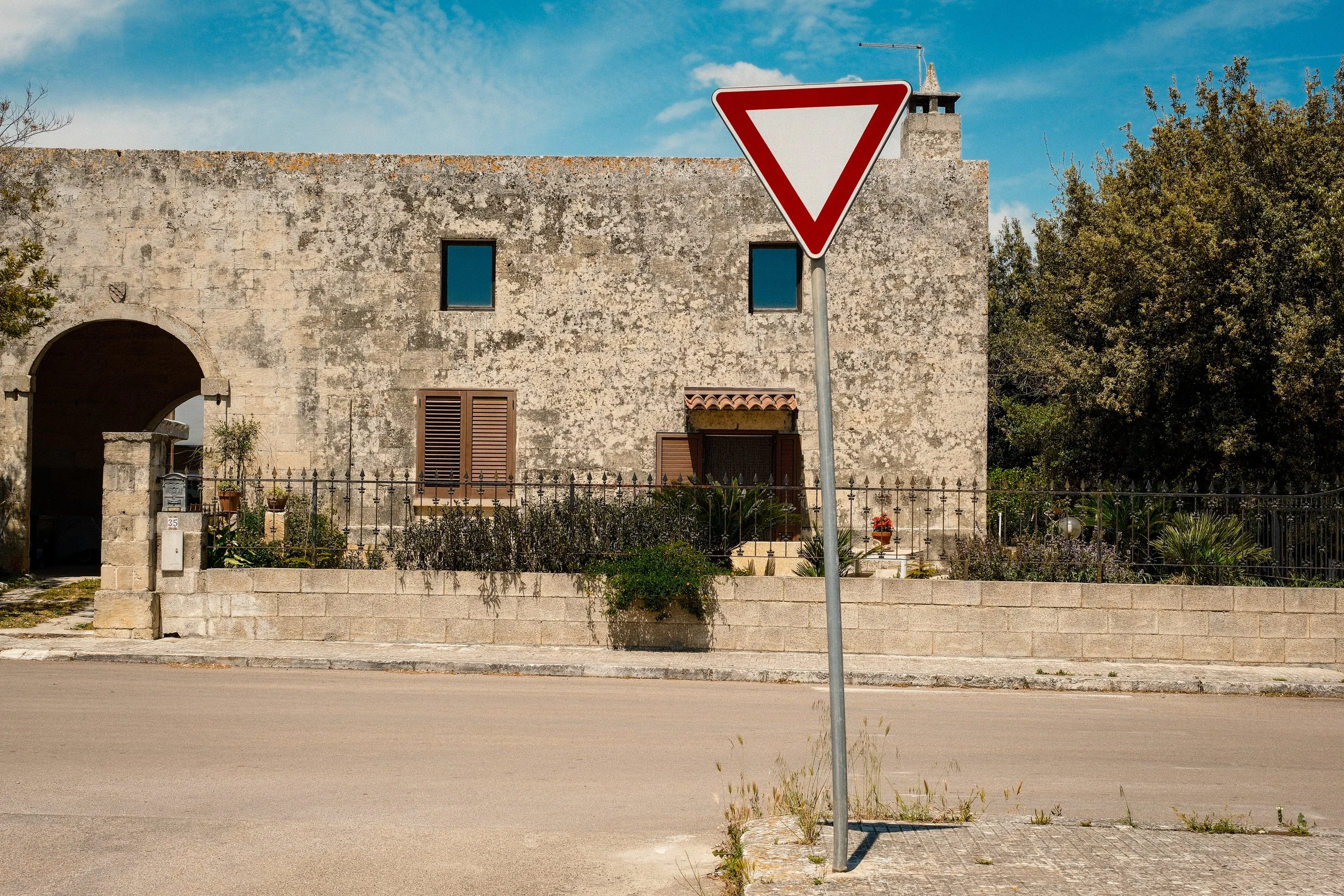 A stone building with barred windows and brown shutters, a small garden with plants and flowers in front, a sidewalk, and a yield traffic sign in the foreground on a sunny day with blue sky and some clouds.