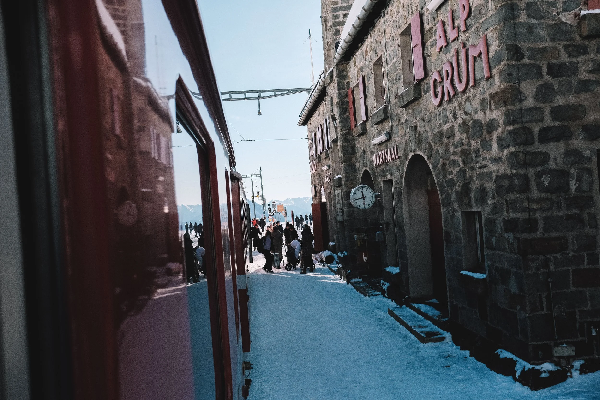 Train station in snowy landscape with red train and stone building labeled 'ALP GRÜM' and 'WARTSAAL', people nearby, mountain backdrop.