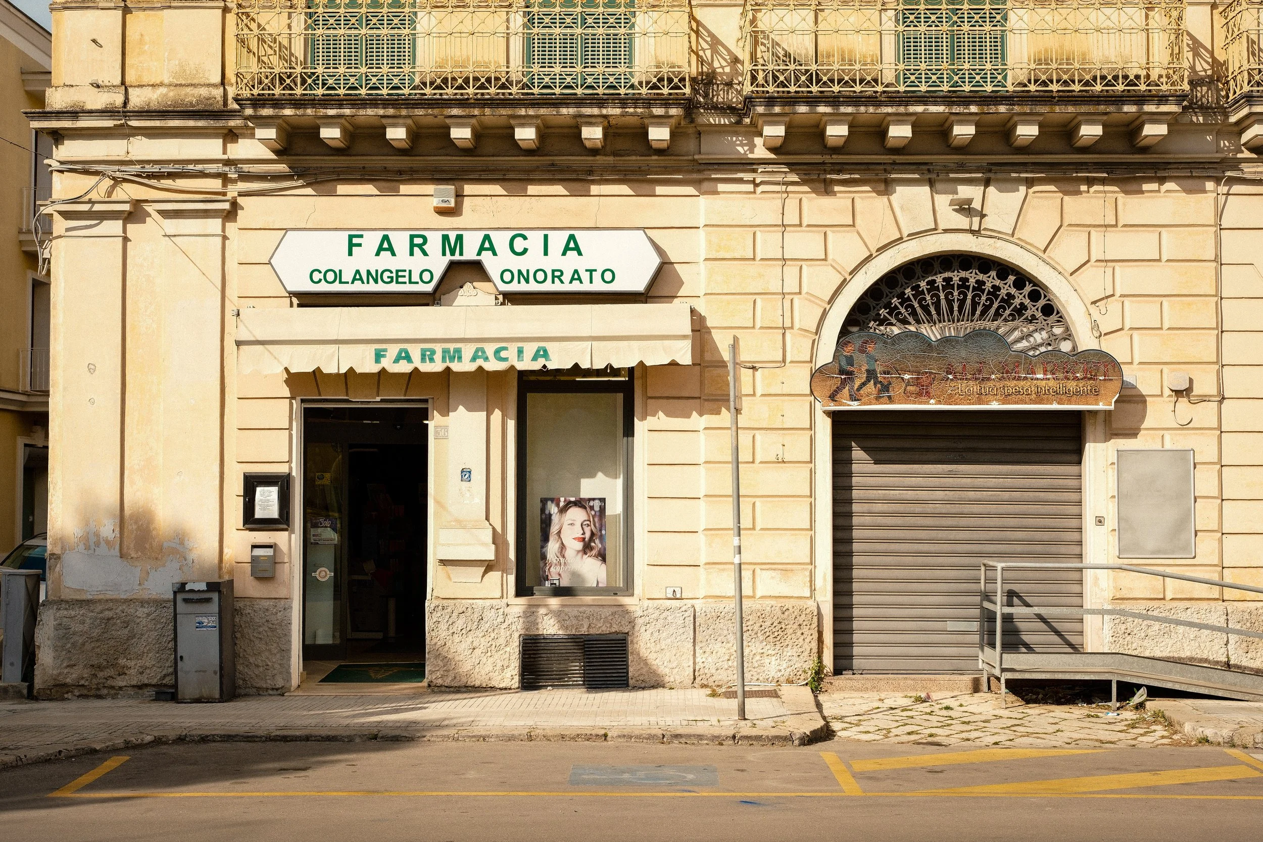 The exterior of a pharmacy with signs reading 'FARMACIA COLANGEL ONARATO' in green letters, a closed metal shutter, a small window with a poster of a woman's face, and a striped awning over the entrance. The building has a beige, somewhat weathered facade with decorative architectural features, and there are parking spaces and a sidewalk in front.