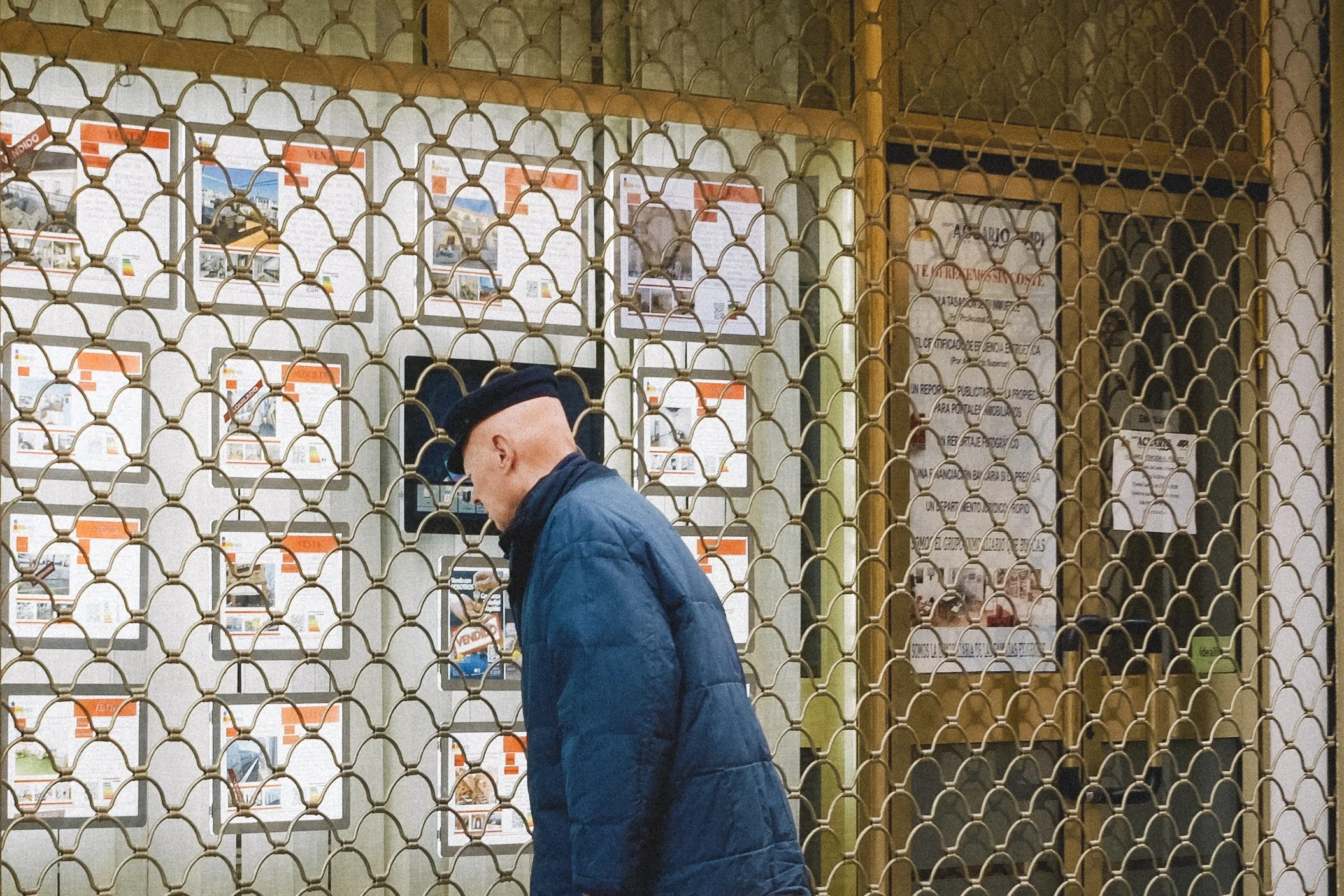 A man in a black beret and blue jacket looking at real estate listings behind a metal gate.