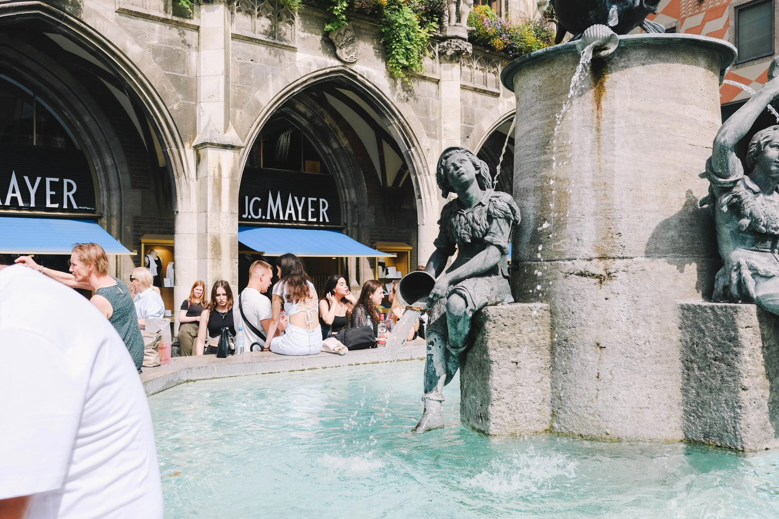 Outdoor scene with a decorative fountain featuring statues, surrounded by people sitting and conversing near the water. The backdrop includes a building with arched windows and a sign reading 'J.G. Mayer.'