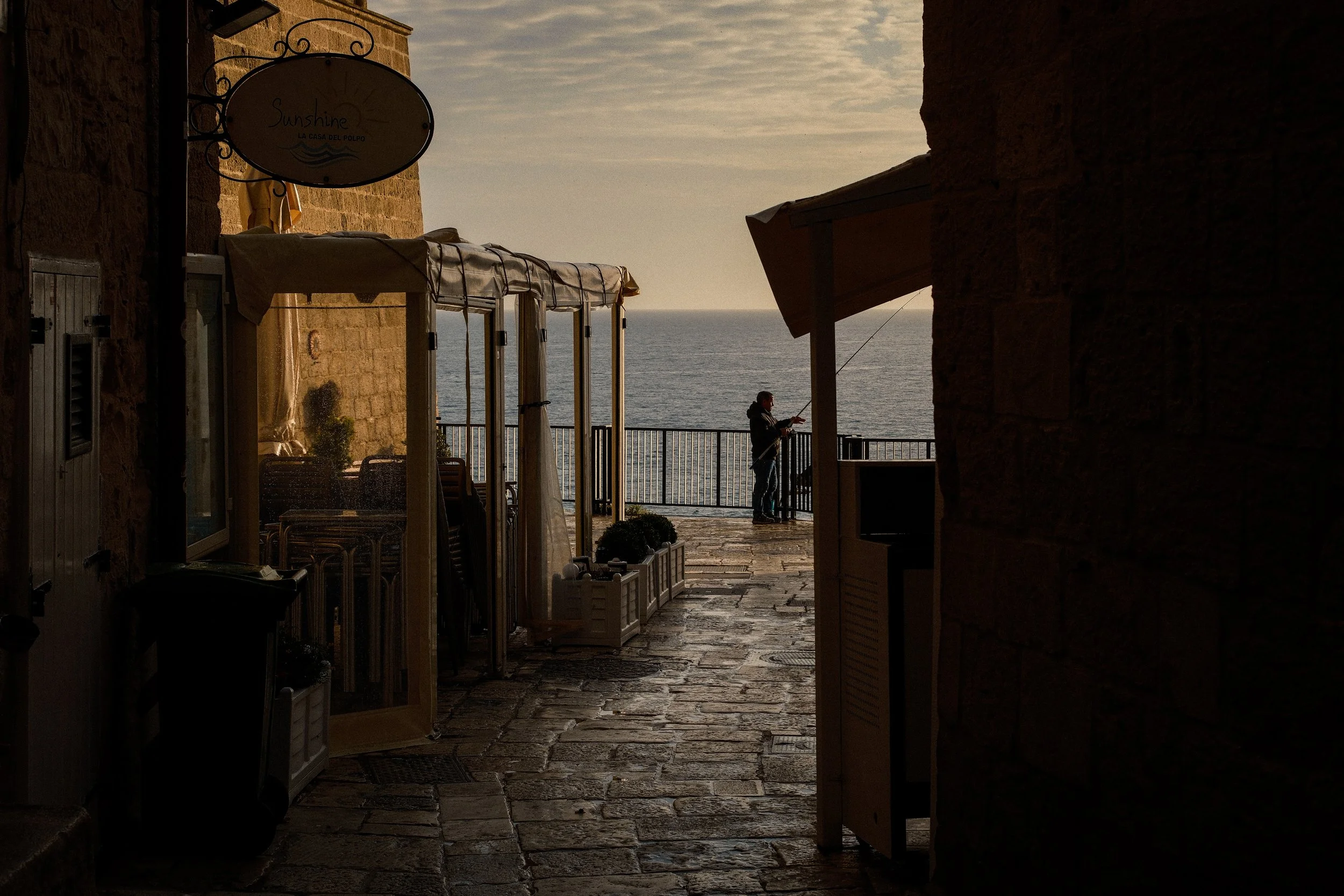 View from a narrow passageway looking out to the ocean. On the left, there are closed shutters and a hanging sign that reads 'Sunshine La Casa del Polpo'. A person is fishing near the railing on the right side, silhouetted against the sea and sky. The scene appears to be during sunset or sunrise with warm, soft light.