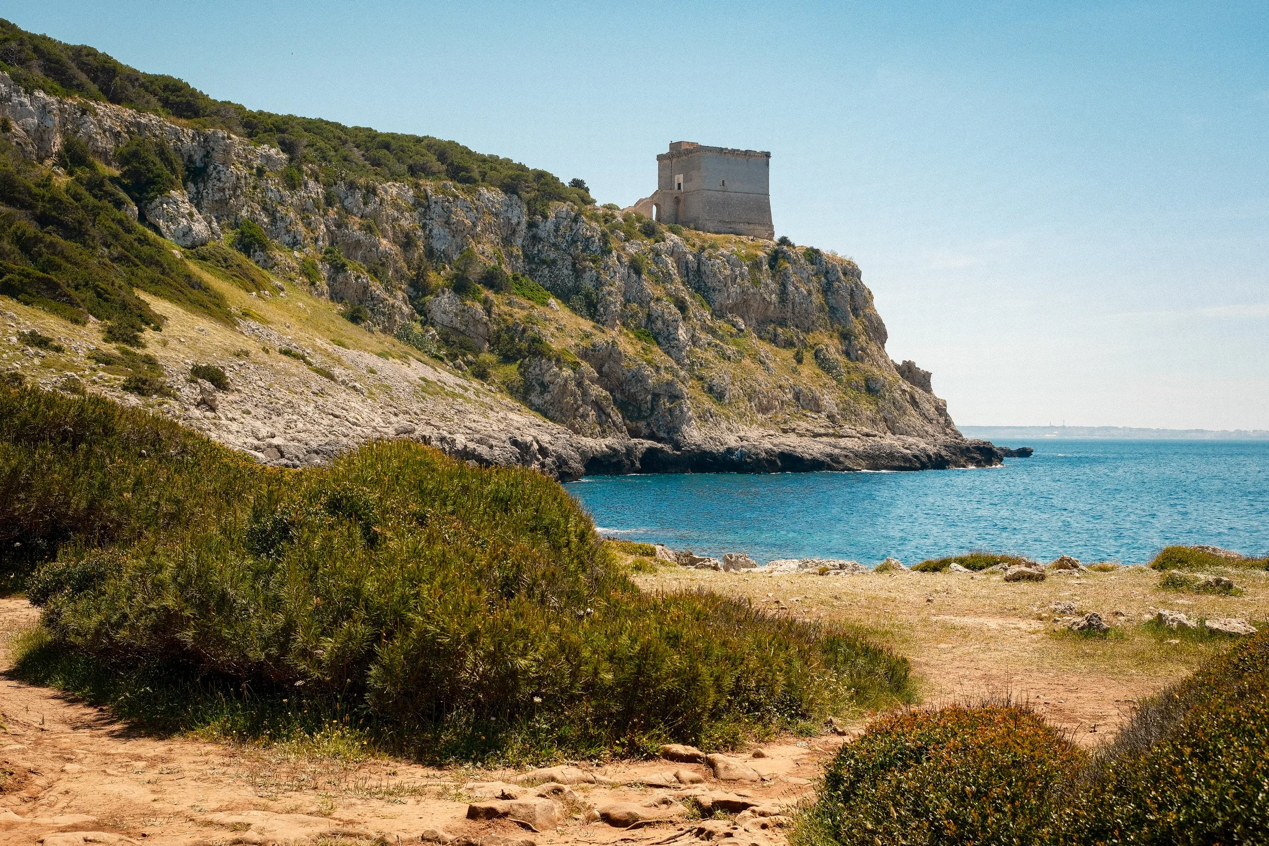 A coastal landscape with a rugged hill covered in green vegetation and a historic stone tower on top, overlooking the blue ocean under a clear sky.