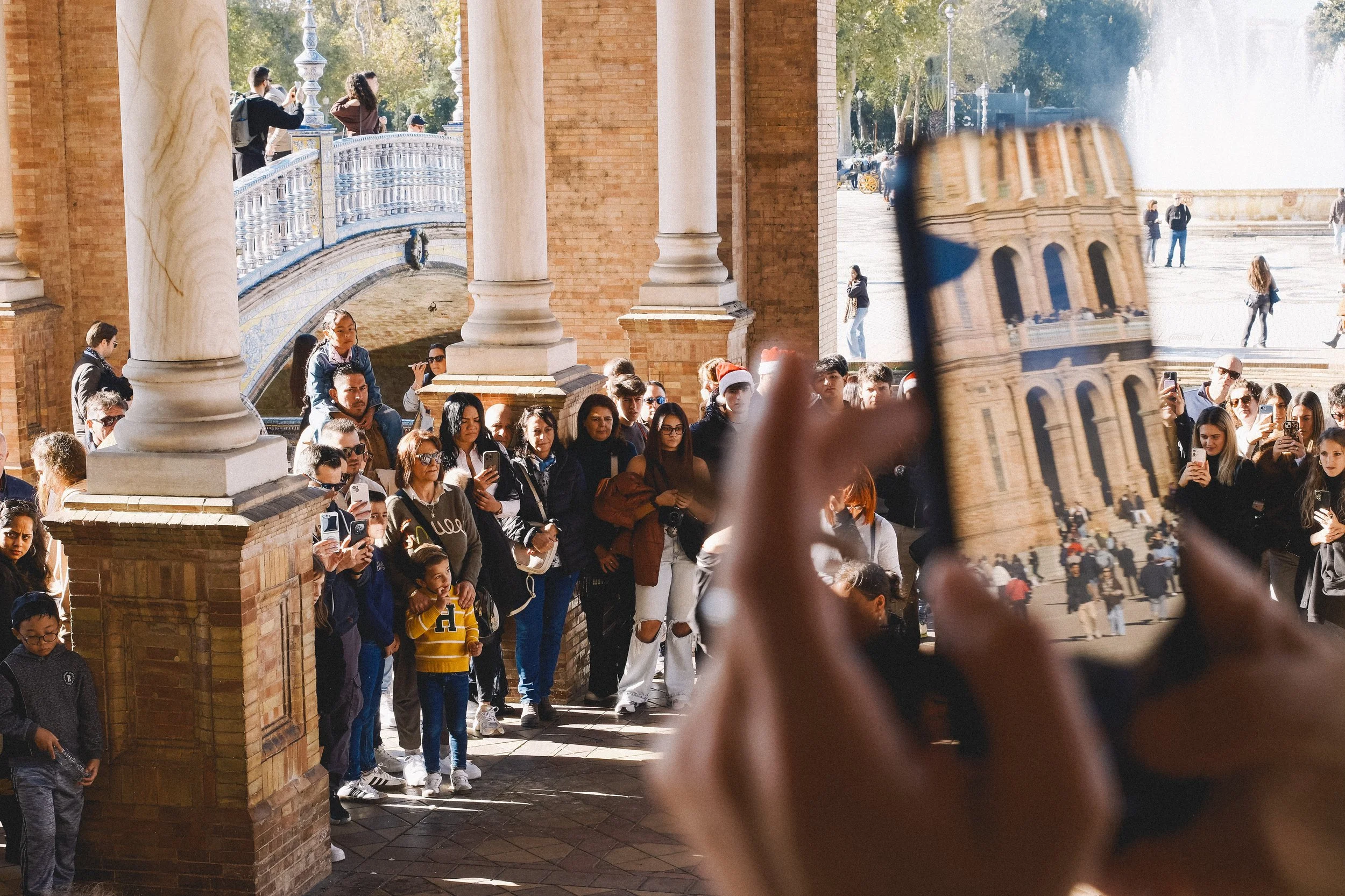 A crowd of tourists gathered at the entrance of a historic building with brick walls and columns, as someone takes a photo with their phone. The background shows a fountain and people walking outside.