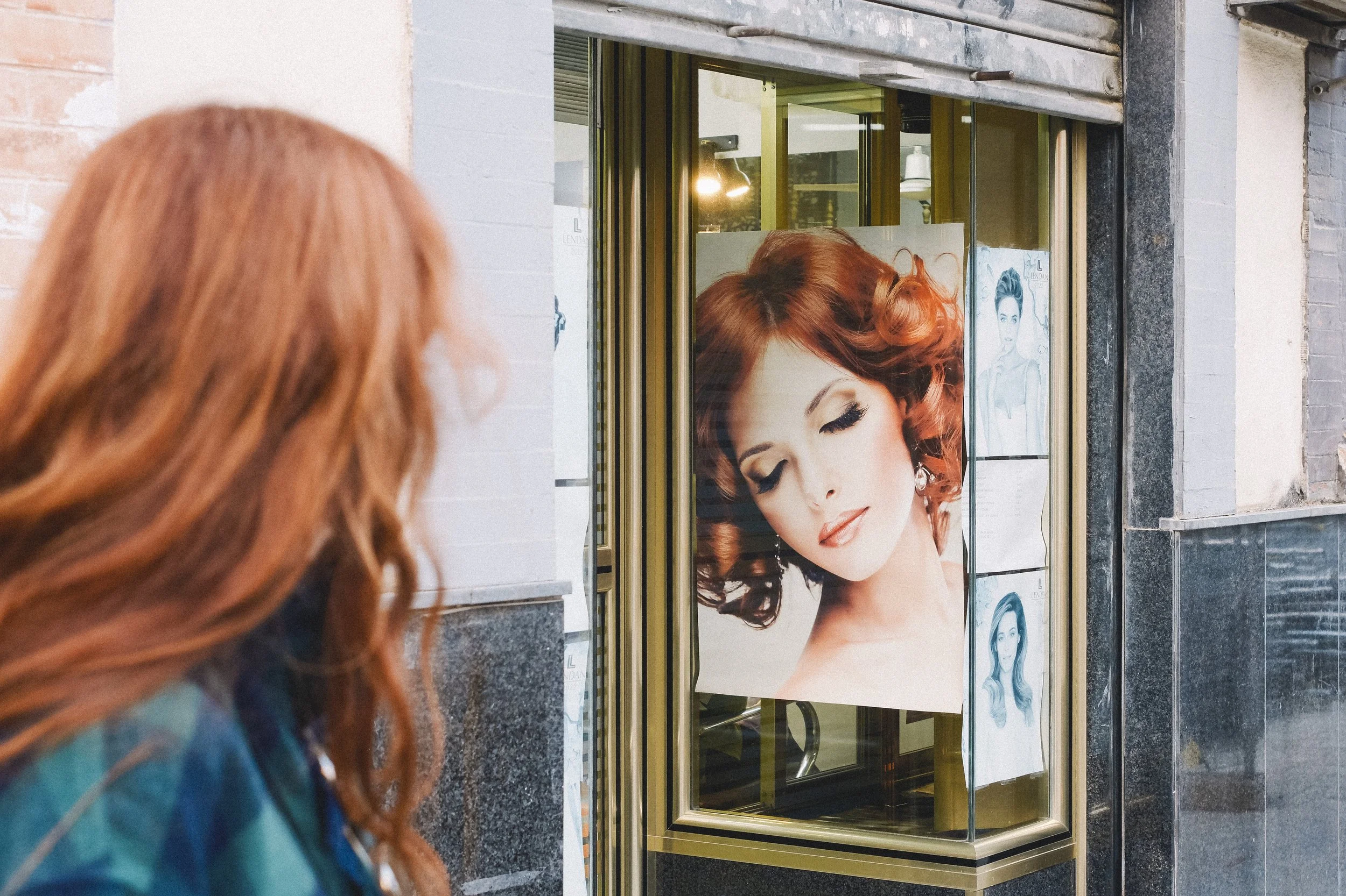 A woman with red hair looking at a large poster of a woman with styled red hair inside a salon window storefront.