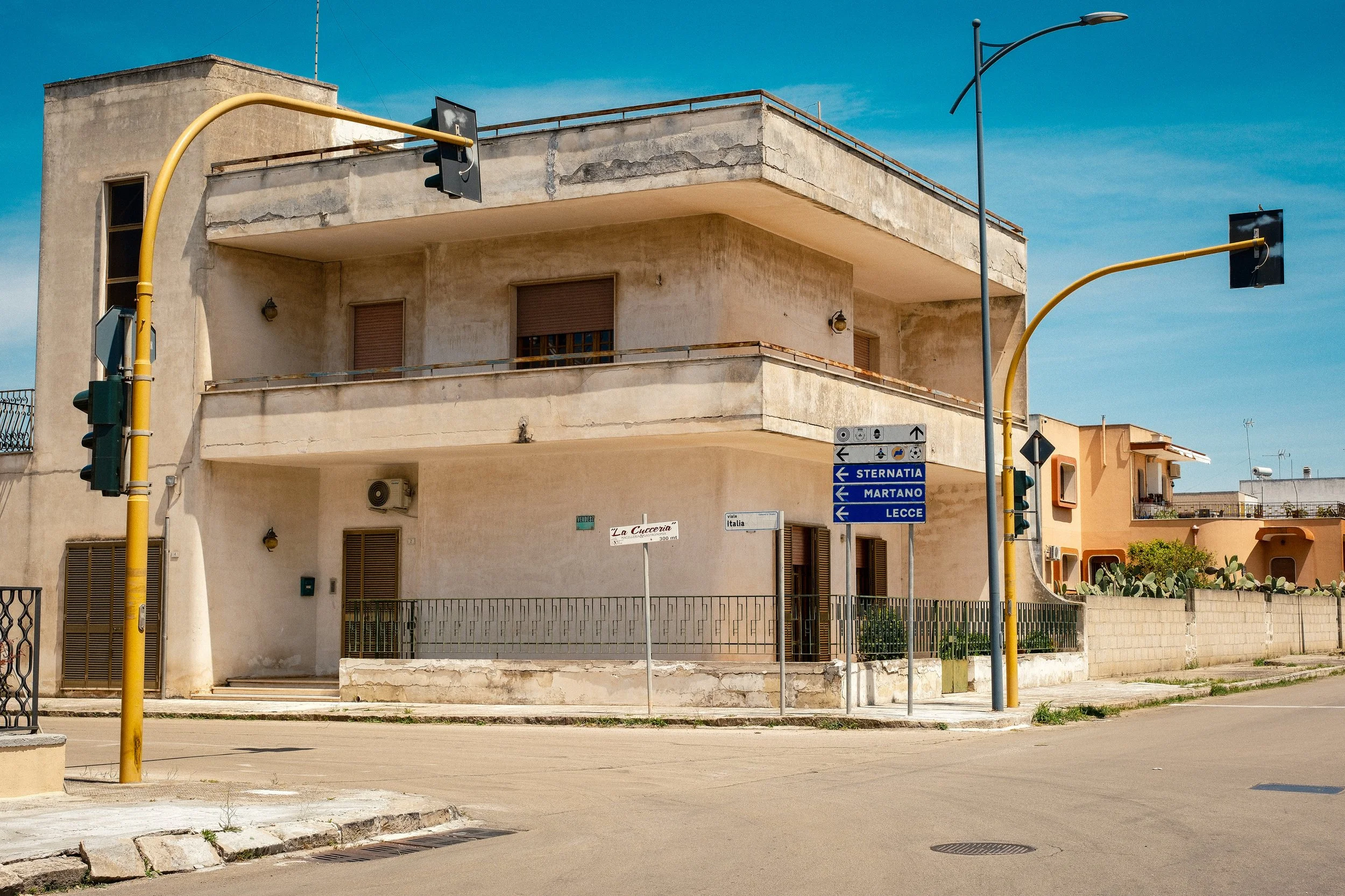 A weathered beige building with two floors, balconies, and closed window shutters. Traffic lights on yellow poles at the street corner, street signs indicating directions to Sernatia, Martano, and Lecce, and a clear blue sky overhead.