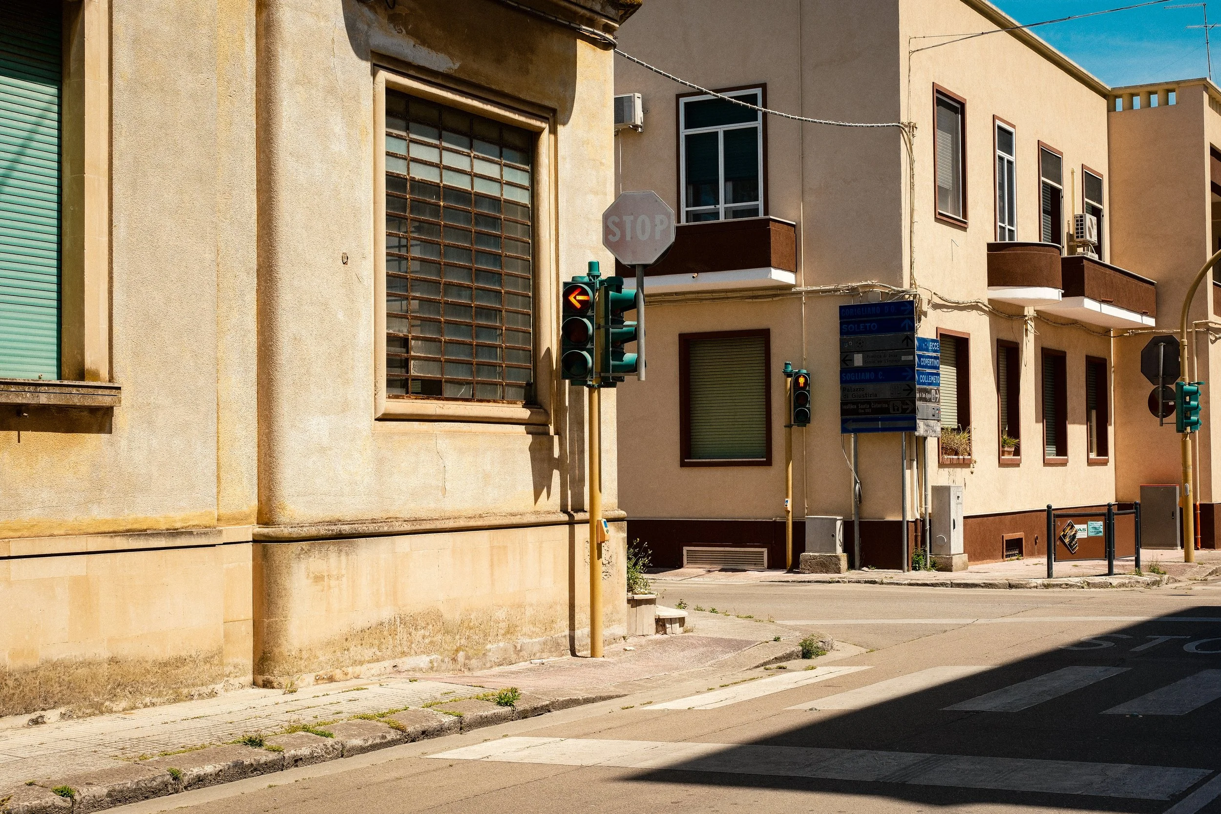 Street scene with a traffic light showing a left arrow and a stop sign in front of beige residential buildings with balconies and shop shutters under a clear blue sky.