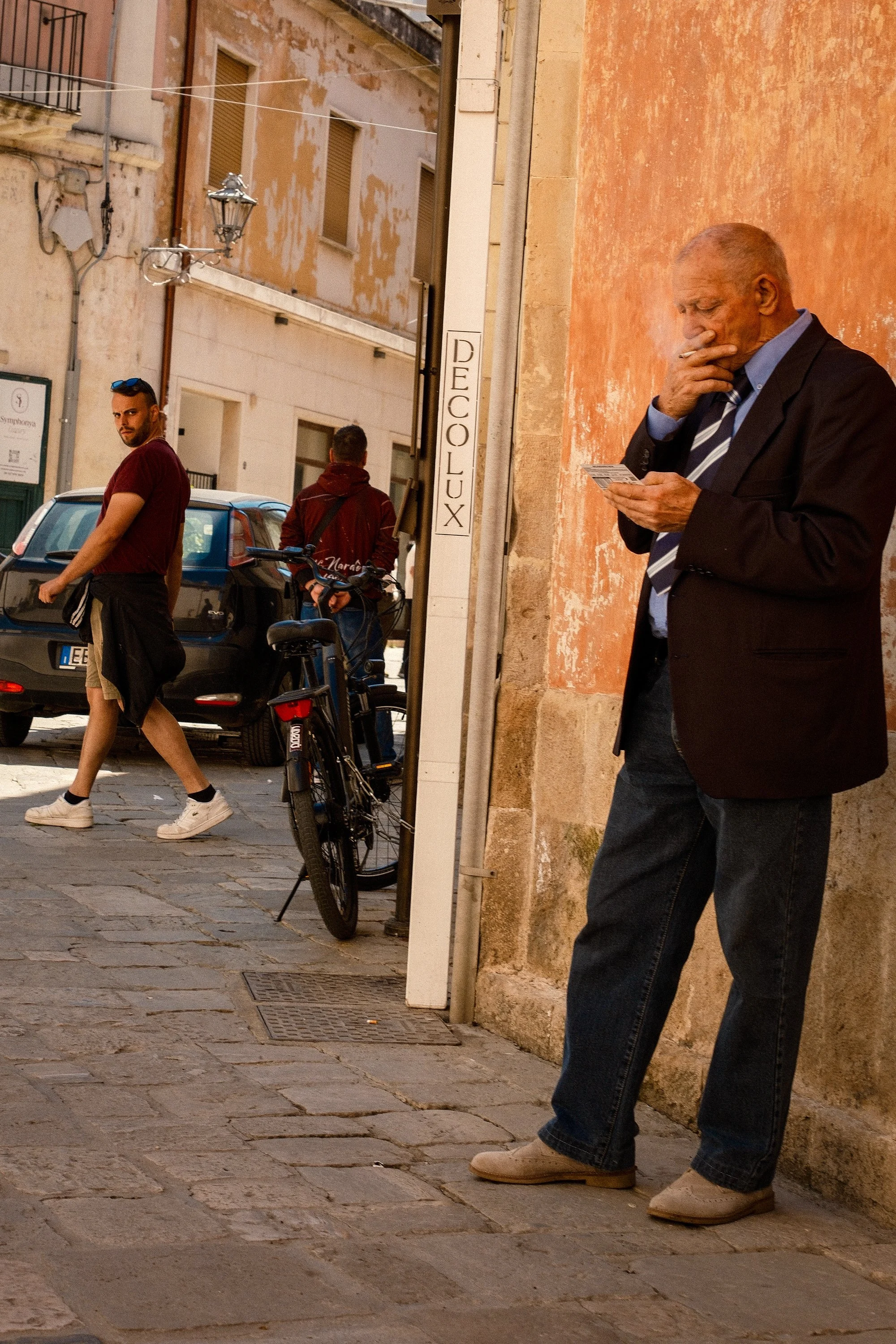 An elderly man with glasses and a striped tie, dressed in a dark blazer and jeans, is standing on a stone sidewalk, smoking and looking at his phone. Behind him, a wall with a weathered orange surface and a sign that says 'DECOLLUX' is visible. In the street, a young man with sunglasses on his head, wearing a maroon t-shirt and khaki shorts, is walking past a parked car and bicycle, with a few other people further in the background.