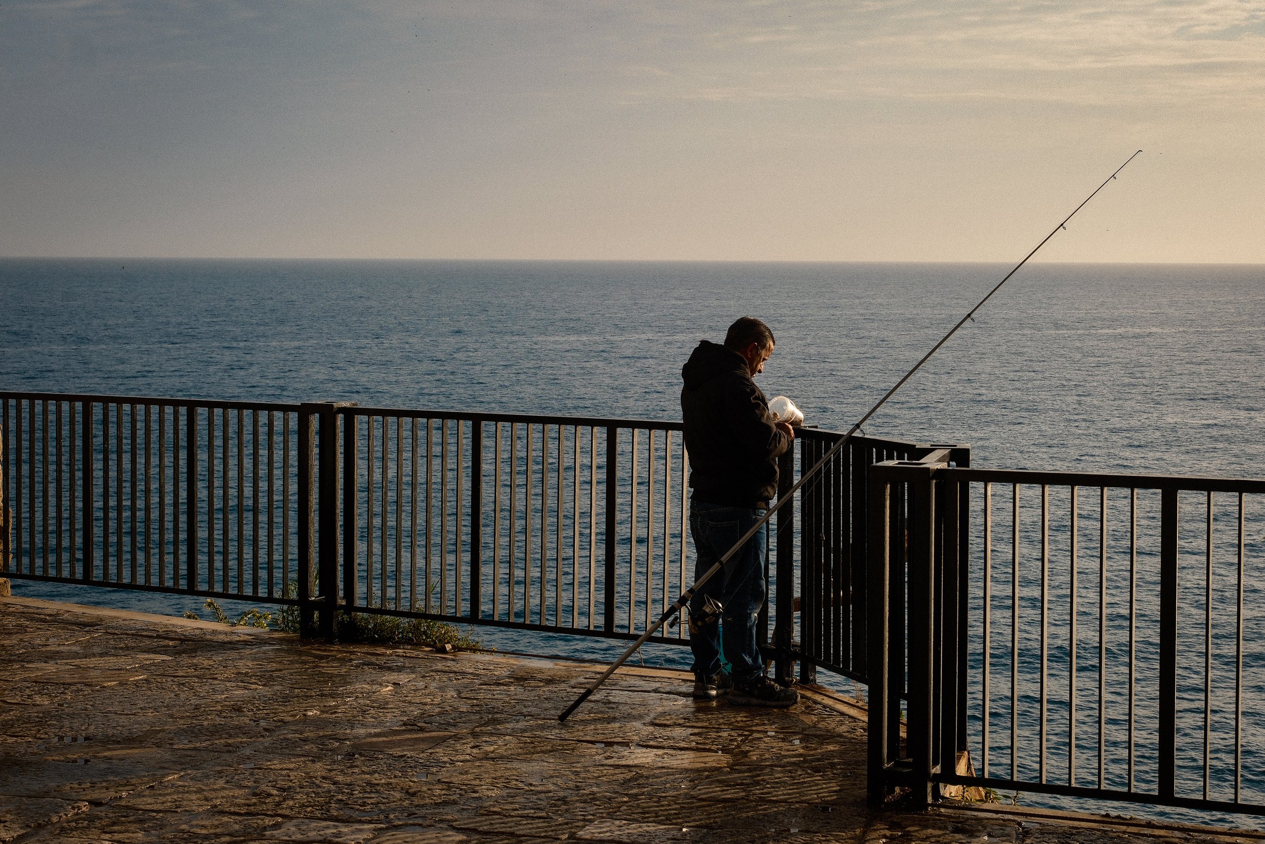 A man fishing off a pier by the ocean during sunset, with a fishing rod cast into the water.