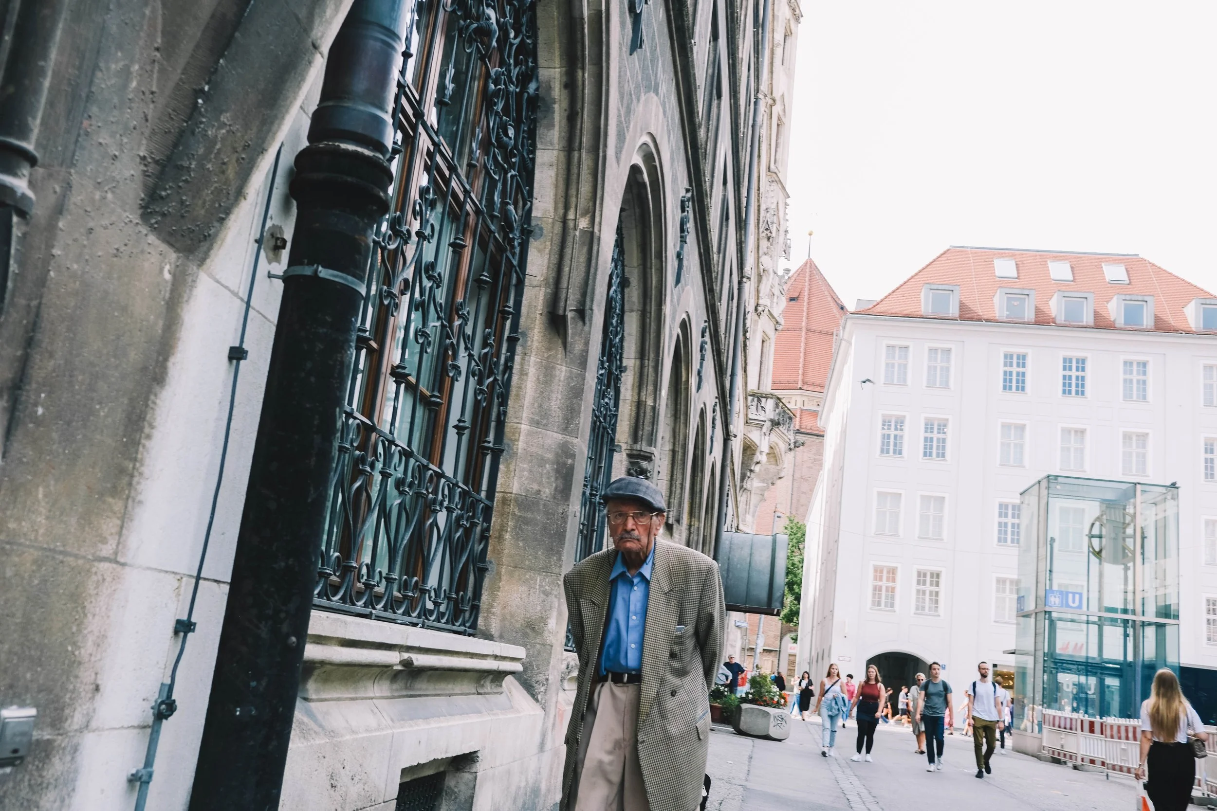 An older man wearing a gray coat and cap walking down a street with historic architecture. The street is lined with buildings with wrought iron details and large windows. People are walking in front of a modern glass structure in the background.