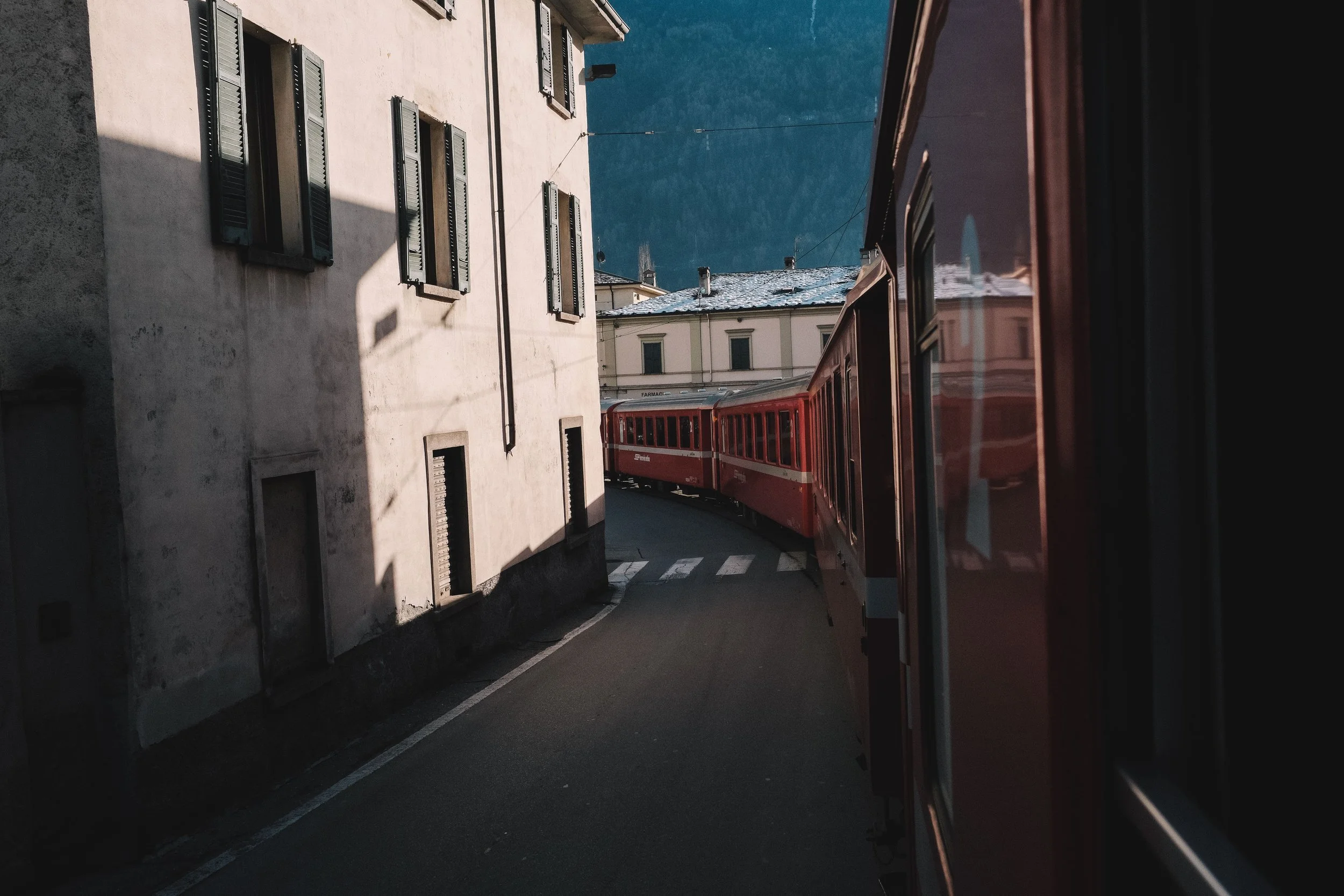 Red train passing through a narrow street with a mountain view in the background.