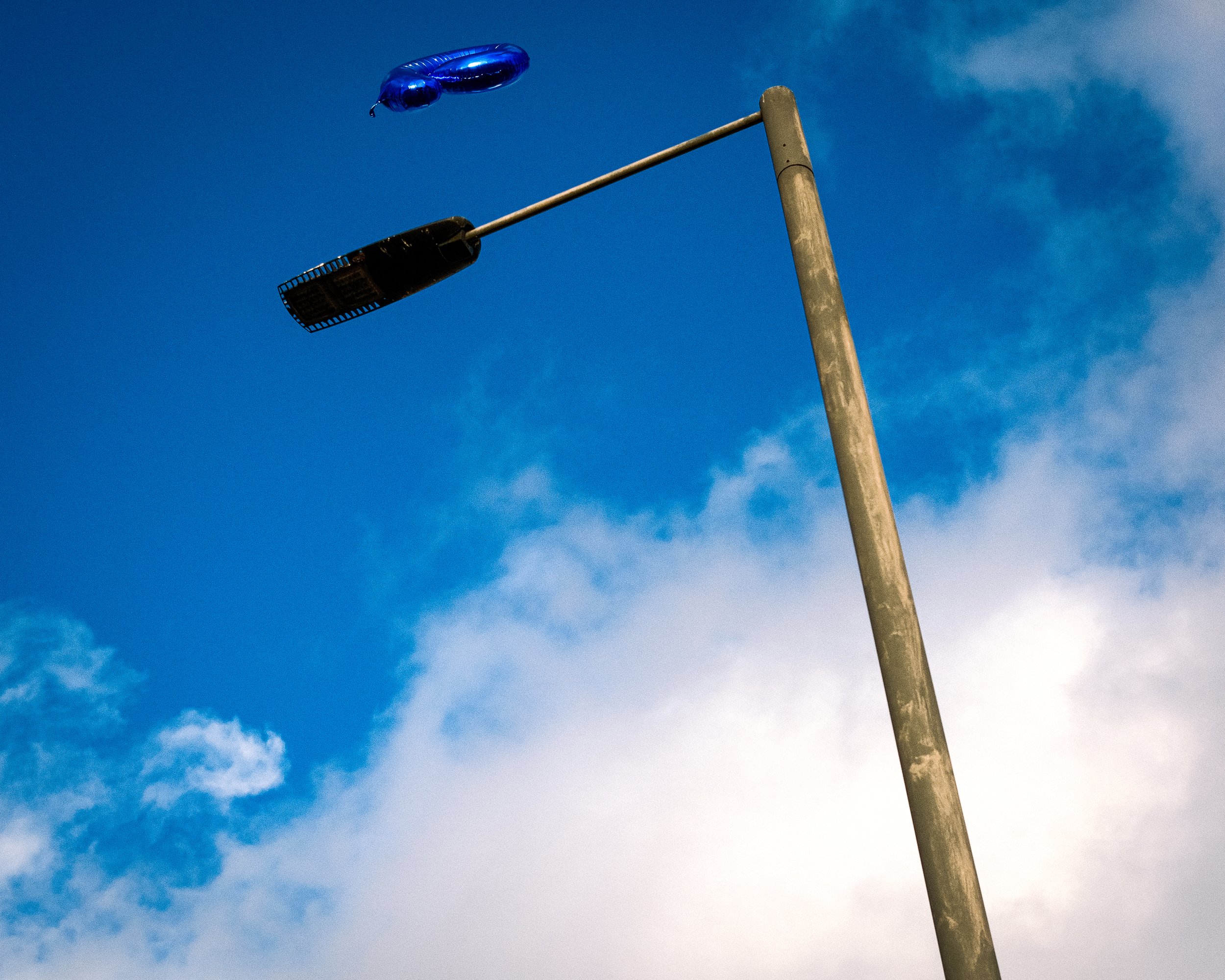 A blue helium balloon floating near a streetlight against a partly cloudy sky.