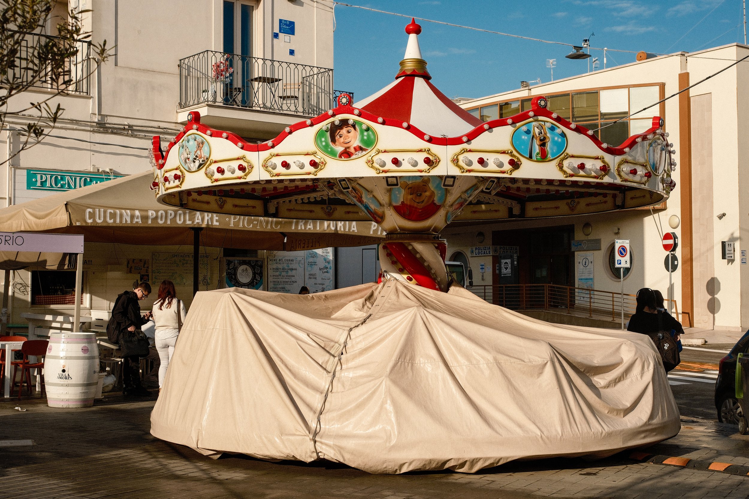 A vintage carousel ride with cartoon character and animal images, parked underneath a beige tent in an outdoor area.