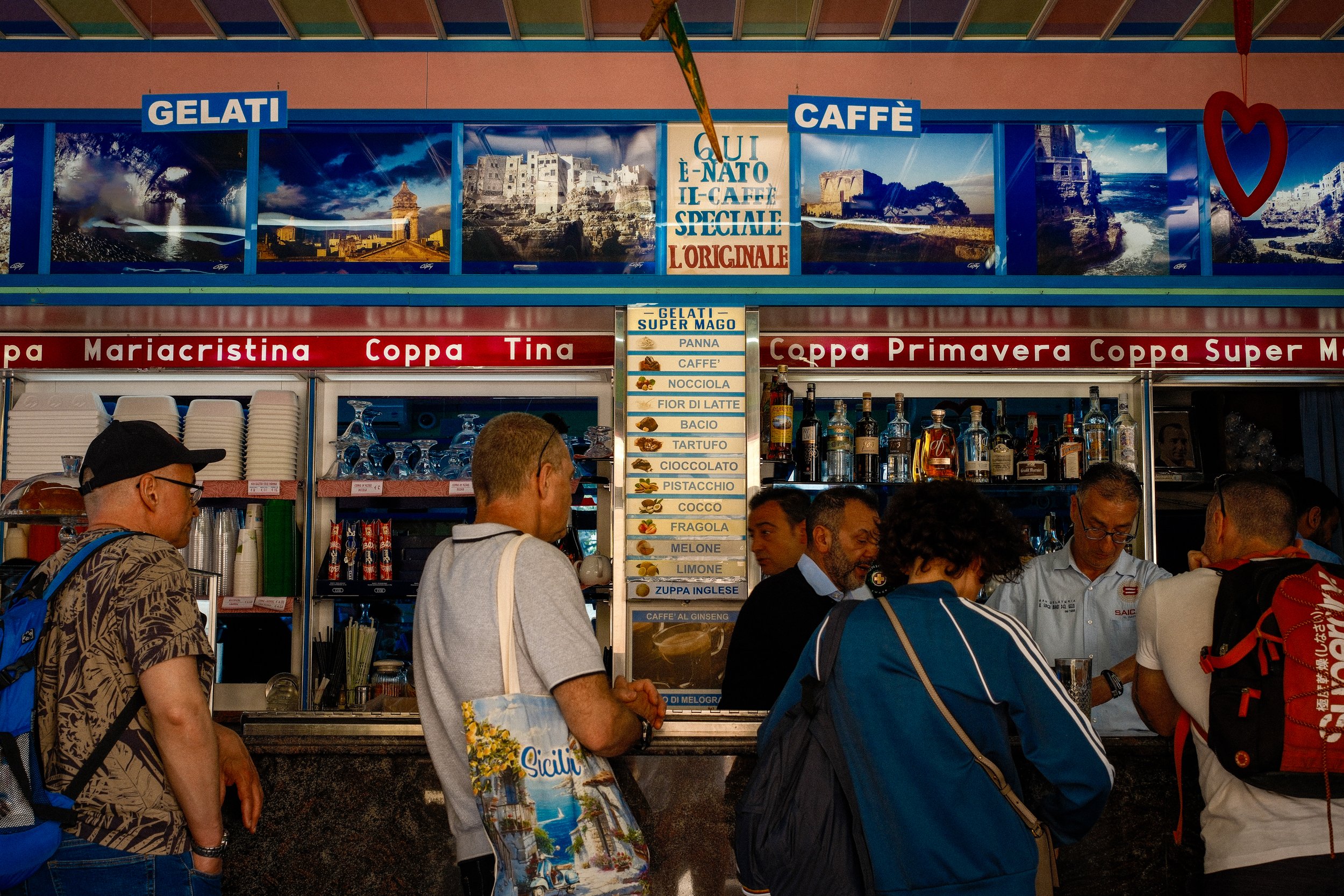 Crowd of people at an outdoor food stand selling ice cream and coffee in Italy, with scenic Italian landscape images on the signage.