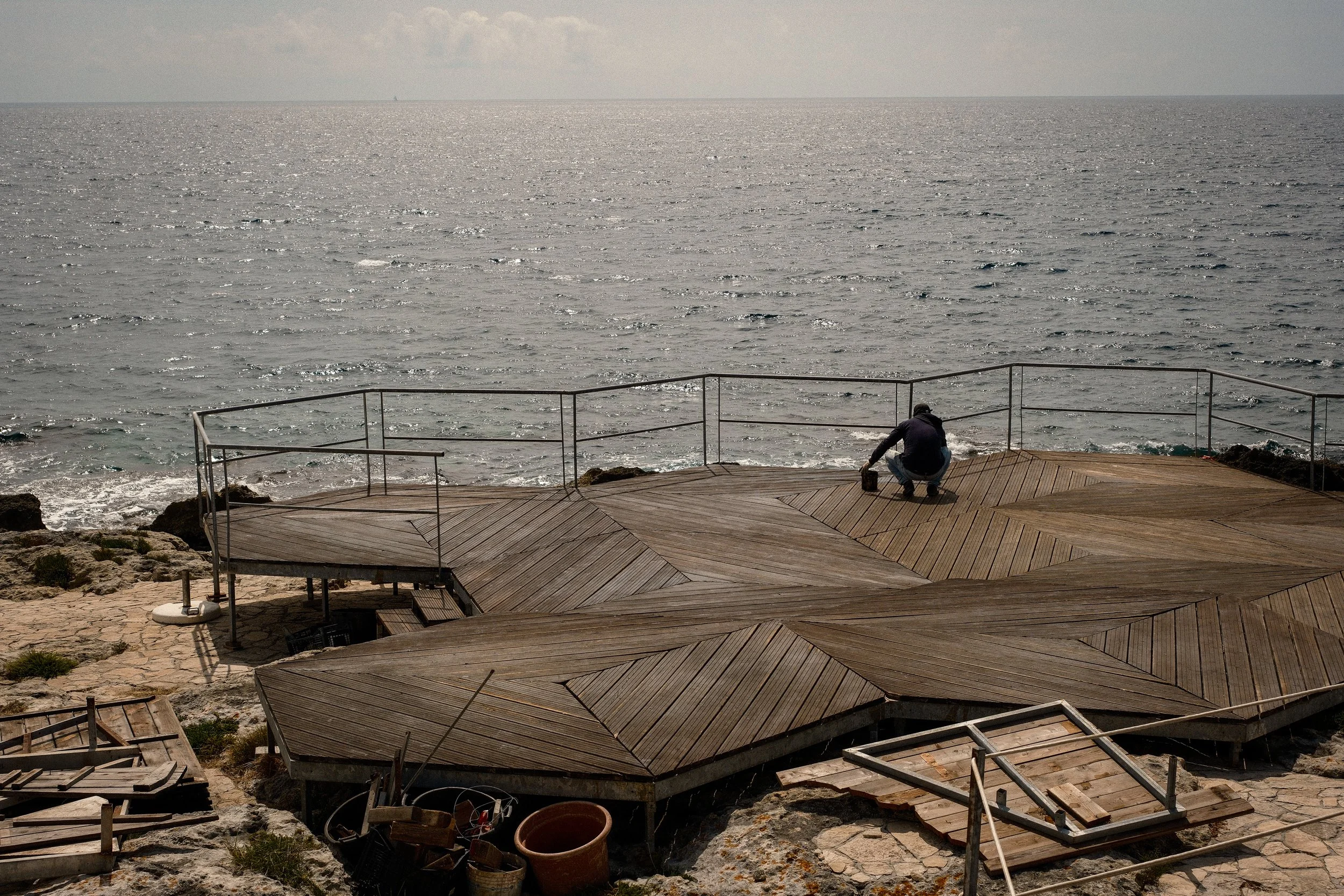 A person crouched down on a wooden deck by the ocean, working with tools near partially assembled structures, with the sea and cloudy sky in the background.