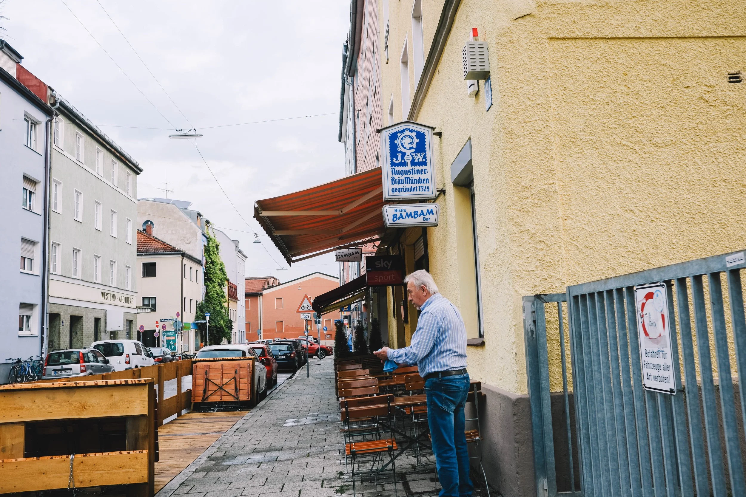 Outdoor scene of a European street with a man standing near an outdoor seating area of a bistro named "BAMBAM." The storefront awning is visible, and there are parked cars along the street. The buildings have various facades and signs in German, indicating a German city setting.