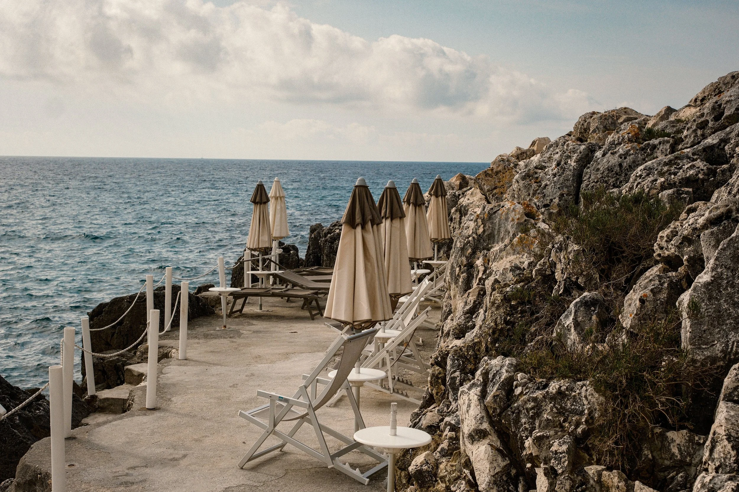 Beachside with a row of closed umbrellas and lounge chairs along a rocky shoreline, overlooking the ocean under a partly cloudy sky.