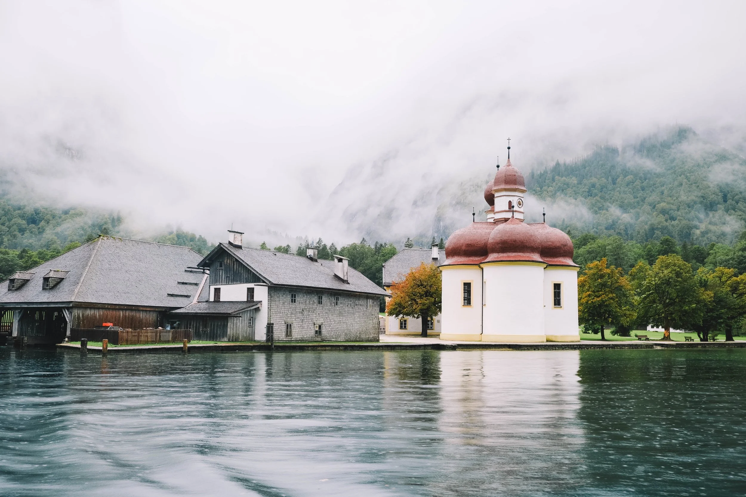 Lakeside building with onion-shaped domes, misty forest backdrop.