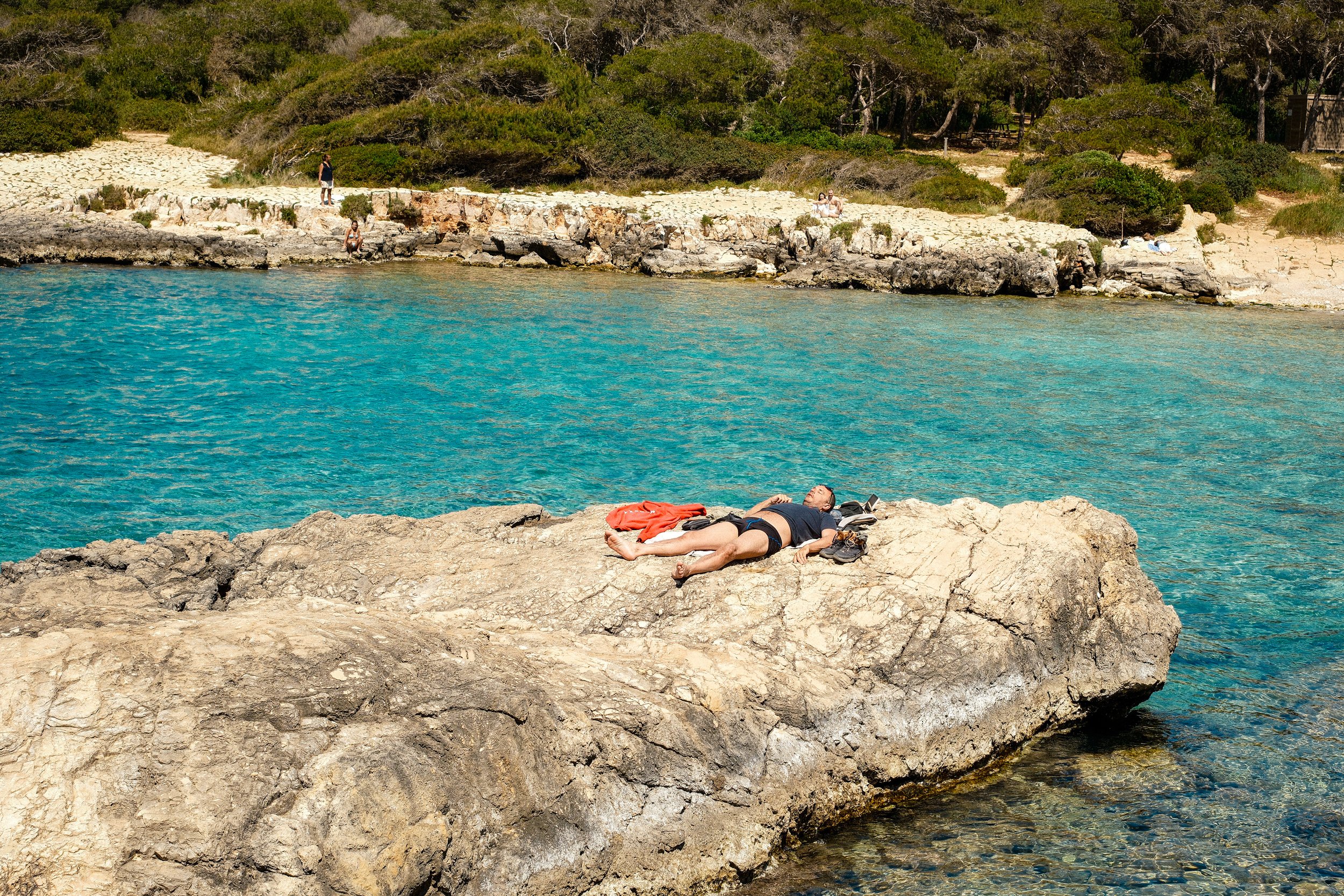 A person lying on a large flat rock near turquoise water, with towels and shoes nearby. In the background, a woman sits on the rocks, and others are walking along the rocky shore near green trees.