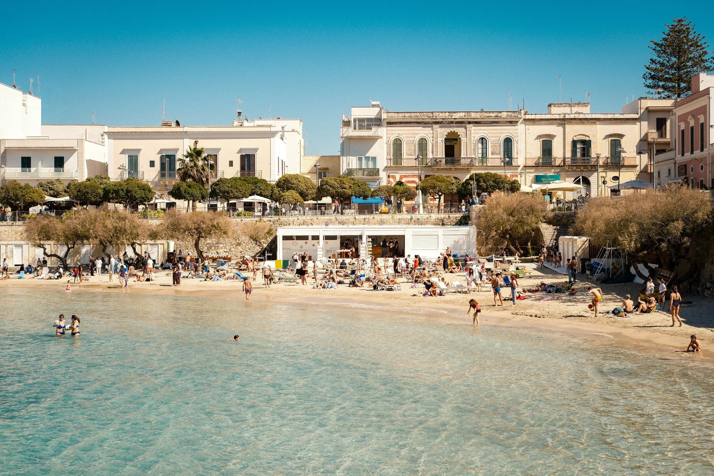 Beach scene with people swimming and relaxing on the sand, overlooking a row of white and pastel-colored buildings with greenery and trees along a promenade.