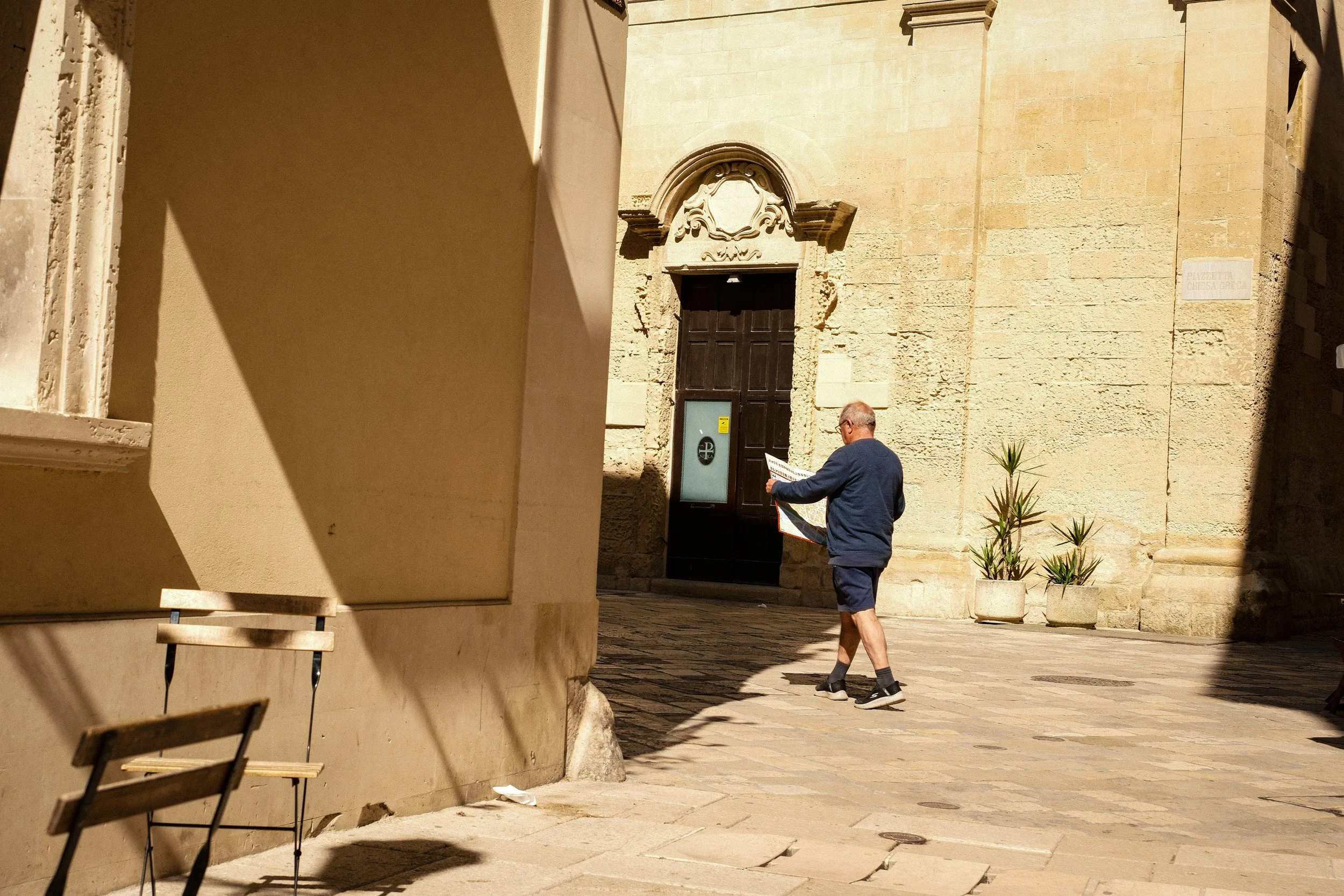 A man reading a newspaper while walking in a European city street with old stone buildings, potted plants, and outdoor furniture.
