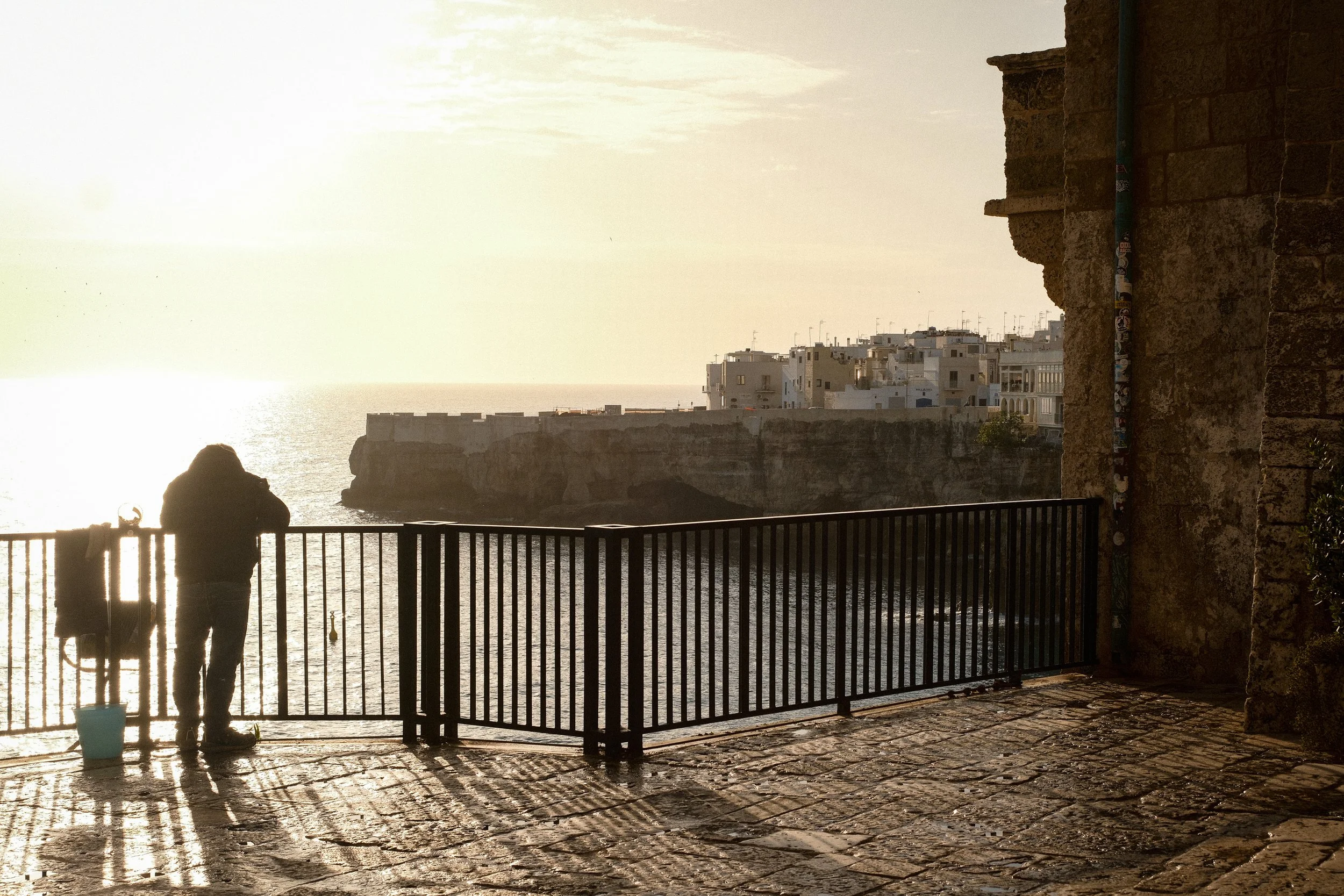 Two people standing and looking at the ocean from a balcony with a metal railing, sunny weather, and buildings in the background.