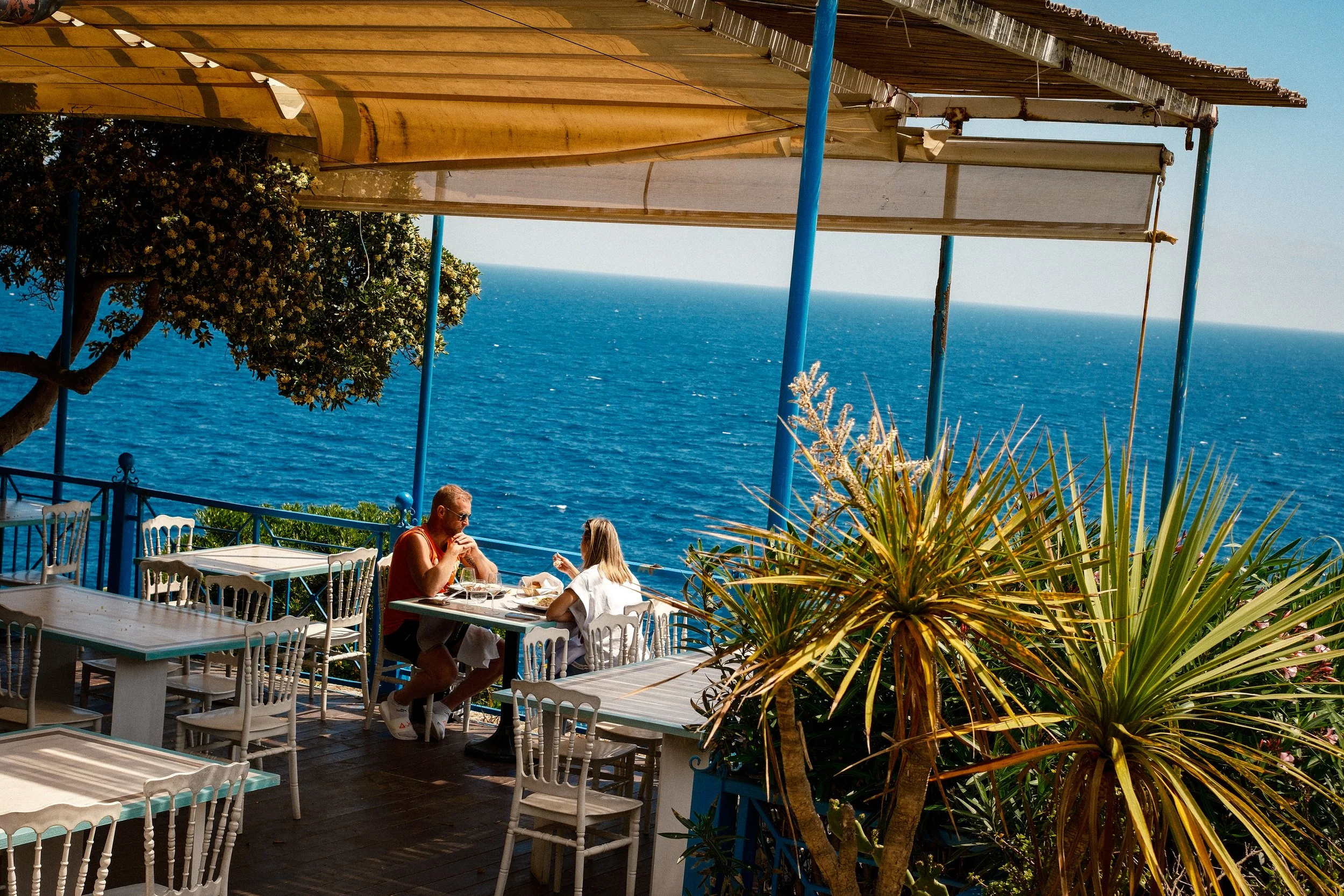 A couple dining outdoors at a restaurant with a view of the ocean, with trees and plants in the foreground and a partly covered patio.