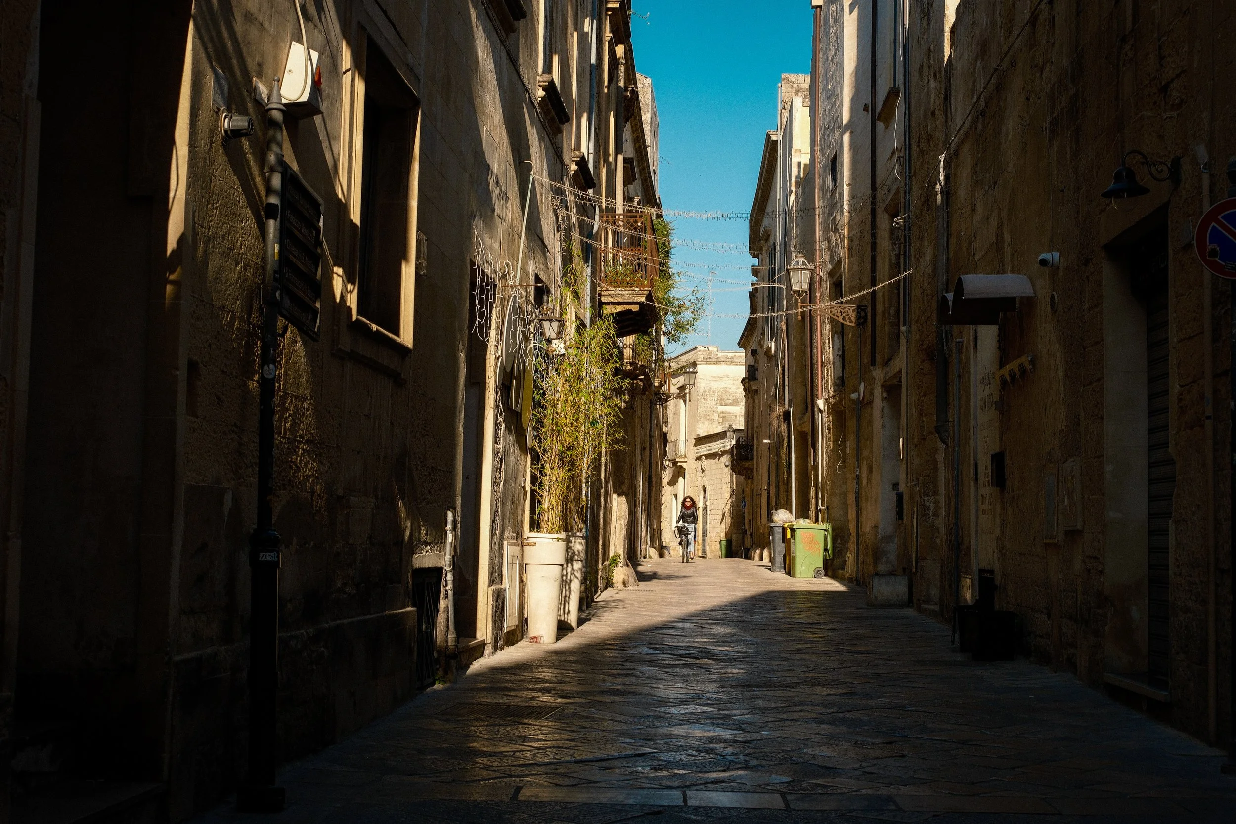 A narrow cobblestone alleyway in an old European city with tall stone buildings on each side. There are some string lights hanging across the alley, and a person riding a bicycle is visible in the distance. The sky is clear and blue.