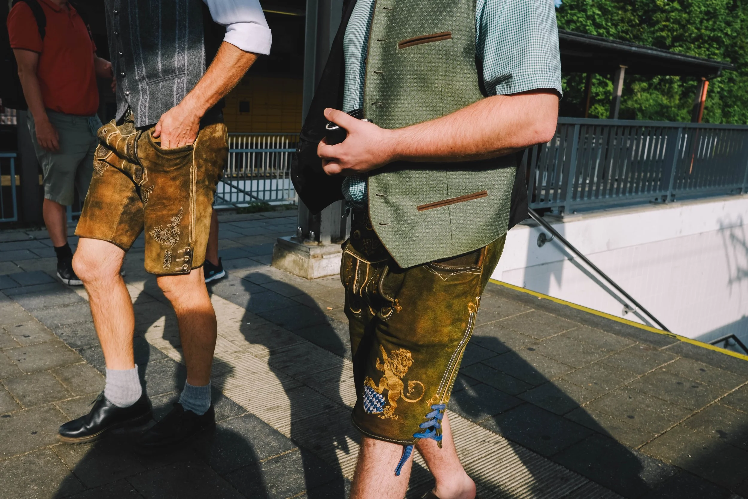 Two men wearing traditional German lederhosen walking outdoors on a sunny day.