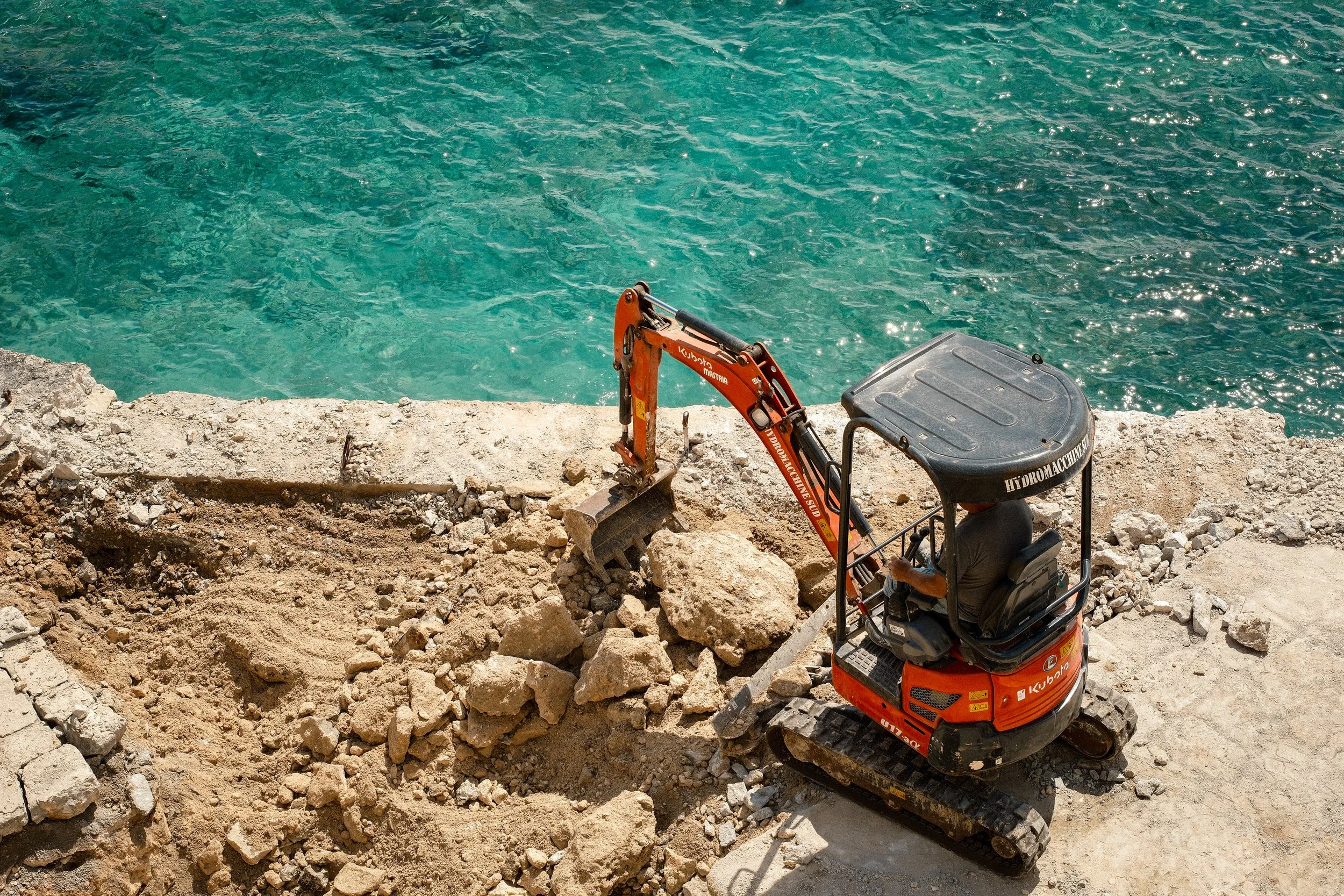 A small orange Kubota mini excavator working on a rocky shoreline near clear blue-green water.