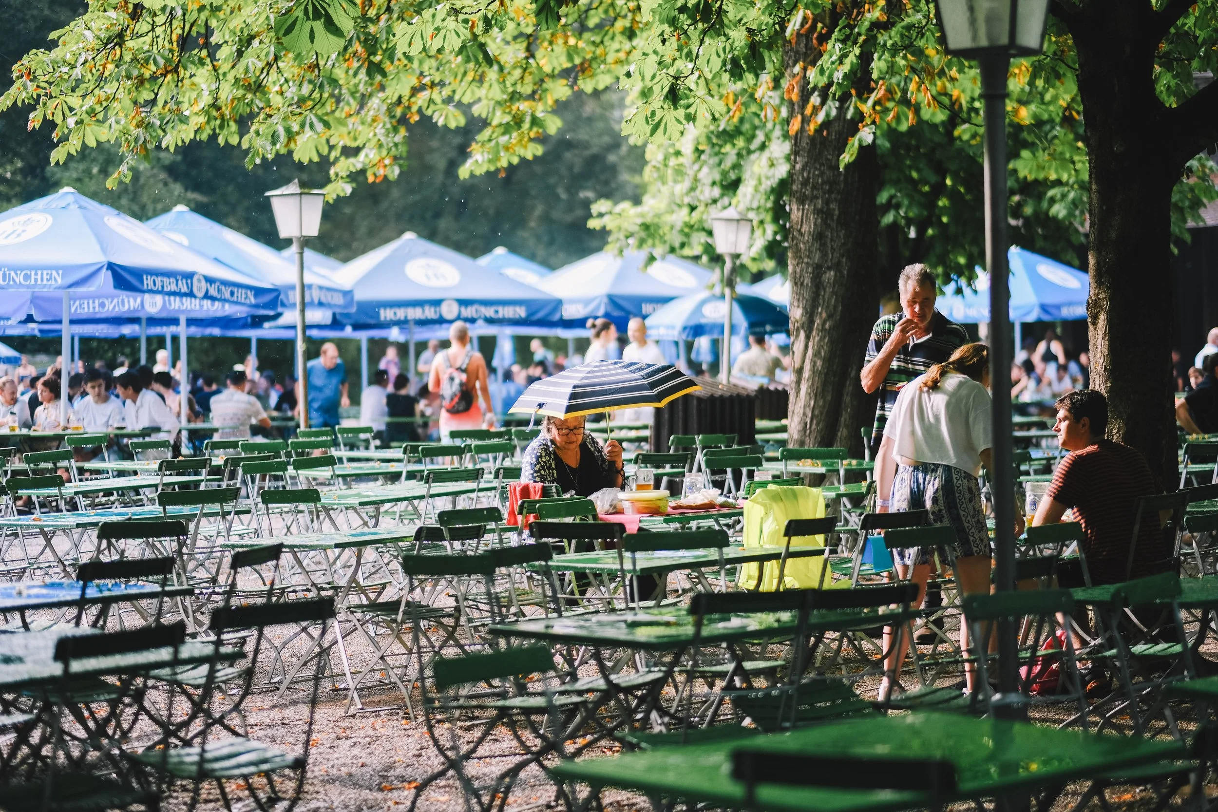 Outdoor beer garden with green tables and chairs, people gathered under blue umbrellas labeled 'Hofbräu München,' surrounded by trees.