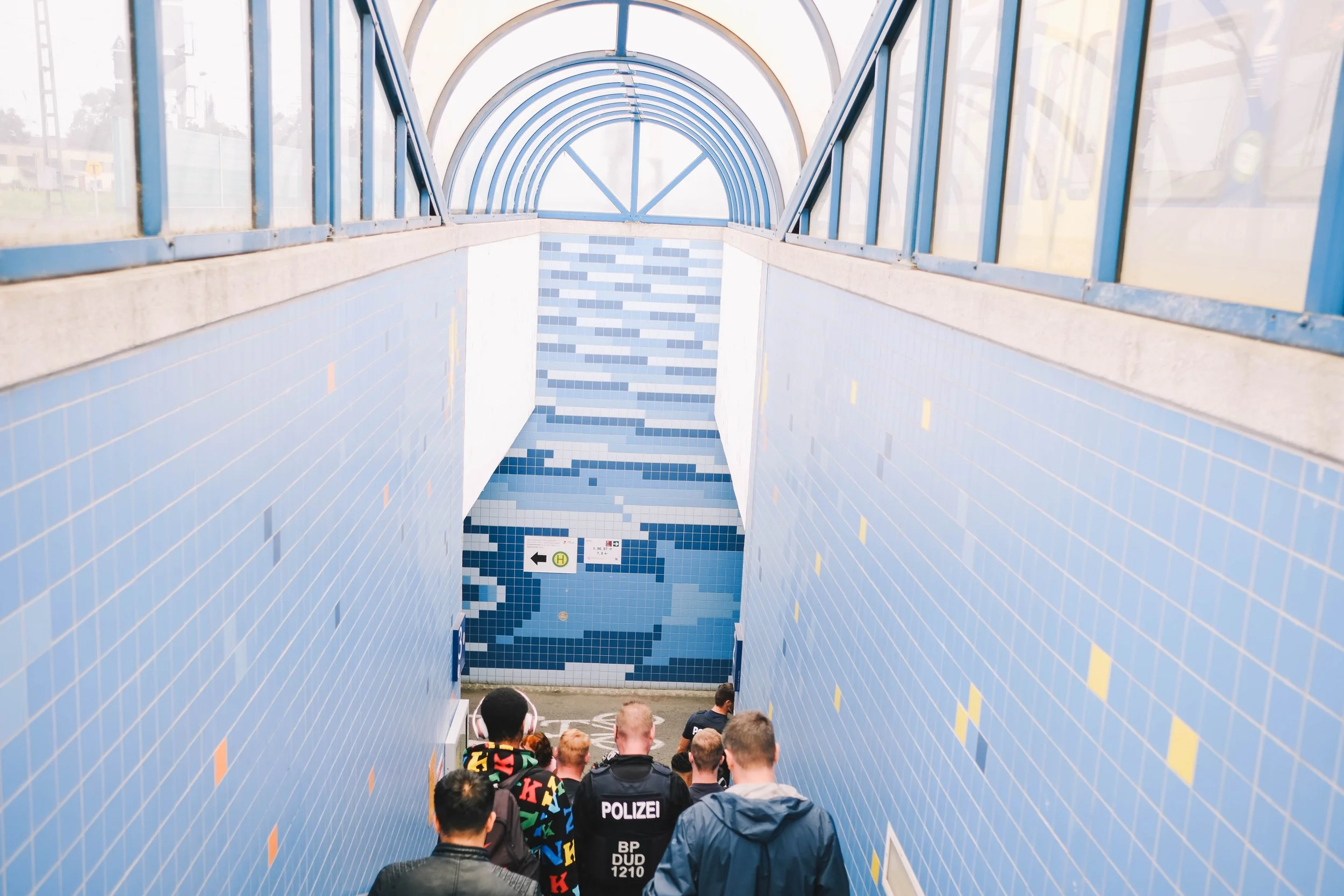 People descending blue-tiled stairs under a glass arched roof with "Polizei" and directional signs visible.