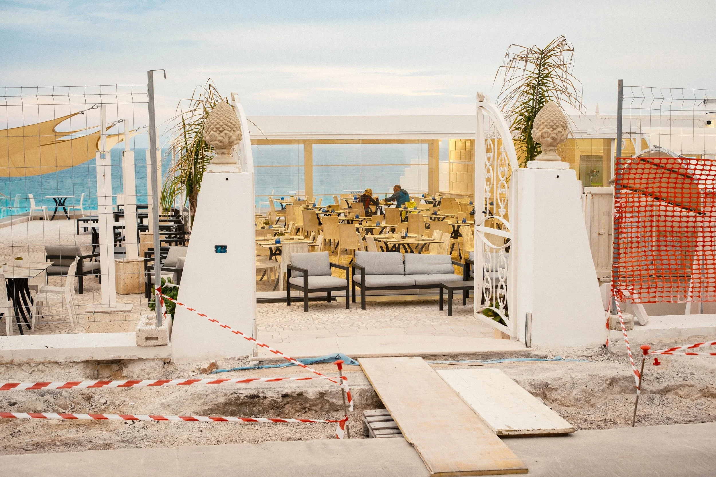 Upscale view of an outdoor seaside restaurant with beige chairs and tables, some tables with blue cups, under a partial awning, viewed through an ornate white gate, with the ocean and sky in the background.