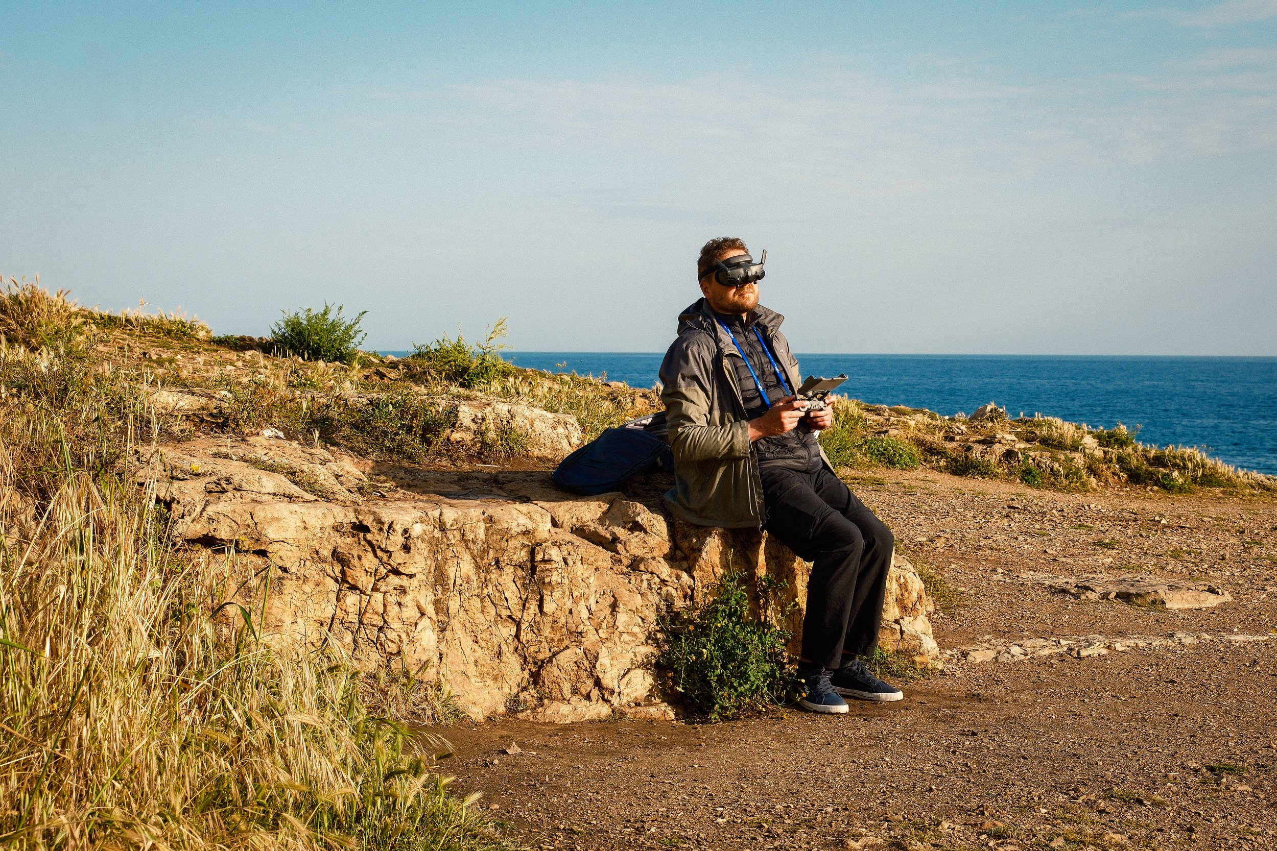 A man with a beard and sunglasses wearing a virtual reality headset sits on a rock by the ocean, holding a remote control, with a backpack on the ground next to him, surrounded by grass and shrubs, under a partly cloudy sky.