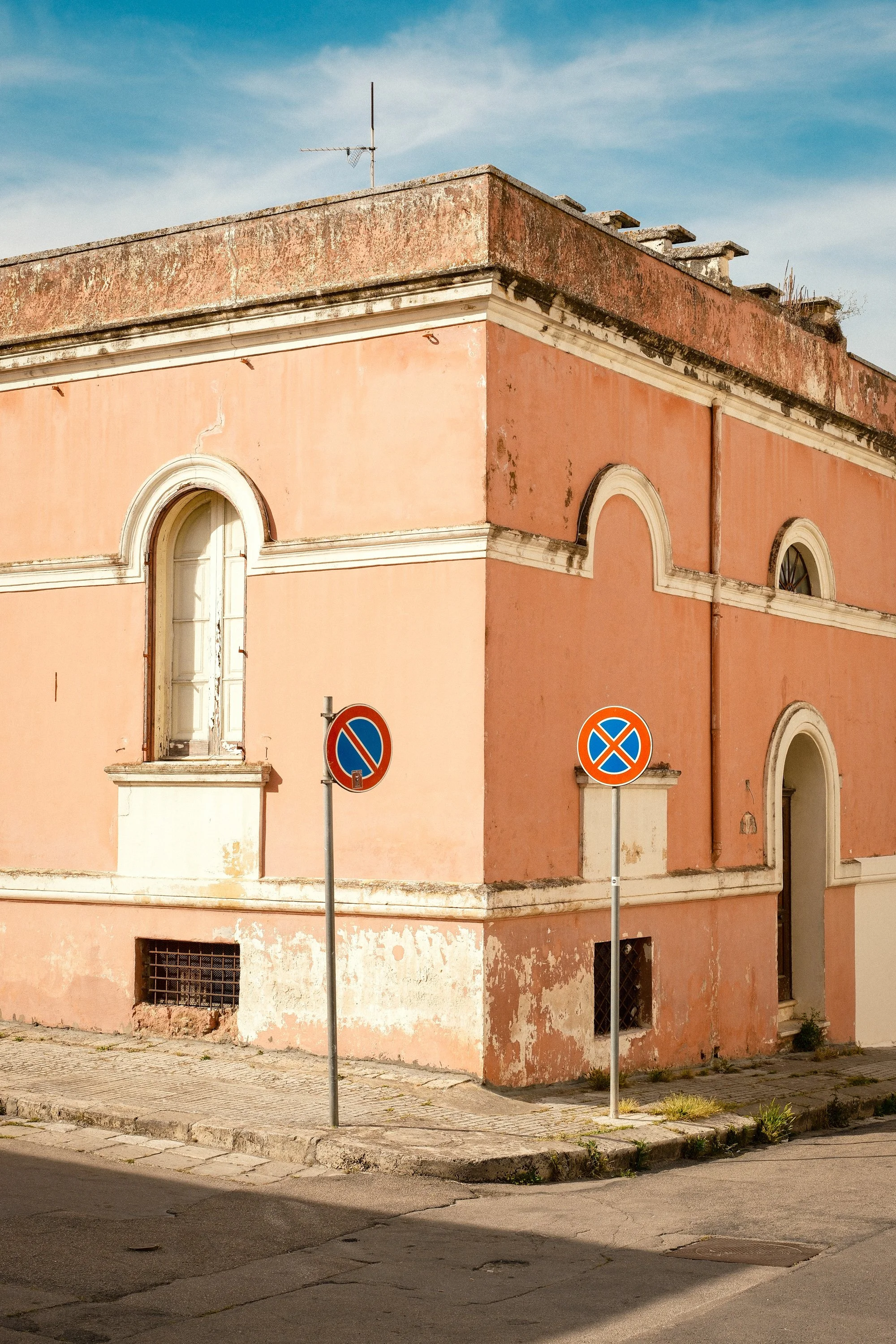 Corner of an old pink building with arched windows and peeling paint, two no parking signs in front, and a street with cracks and patches.