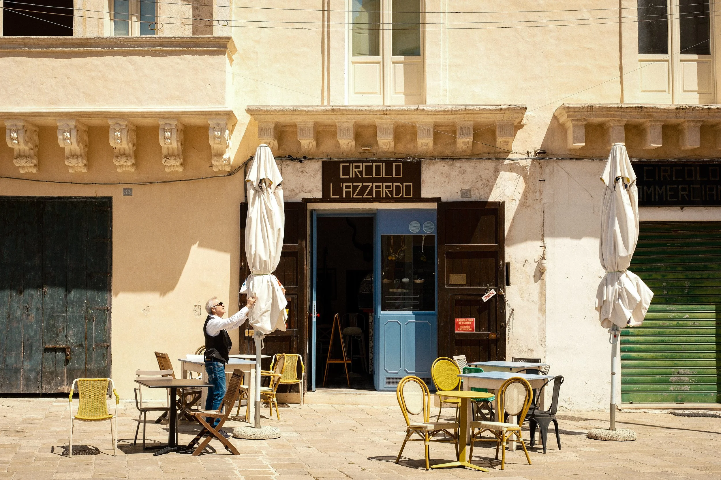 Outdoor cafe with empty tables and chairs, closed umbrellas, and a person setting up an umbrella in front of a building with signs that read Circolo L'azzardo, with a balcony and windows above.