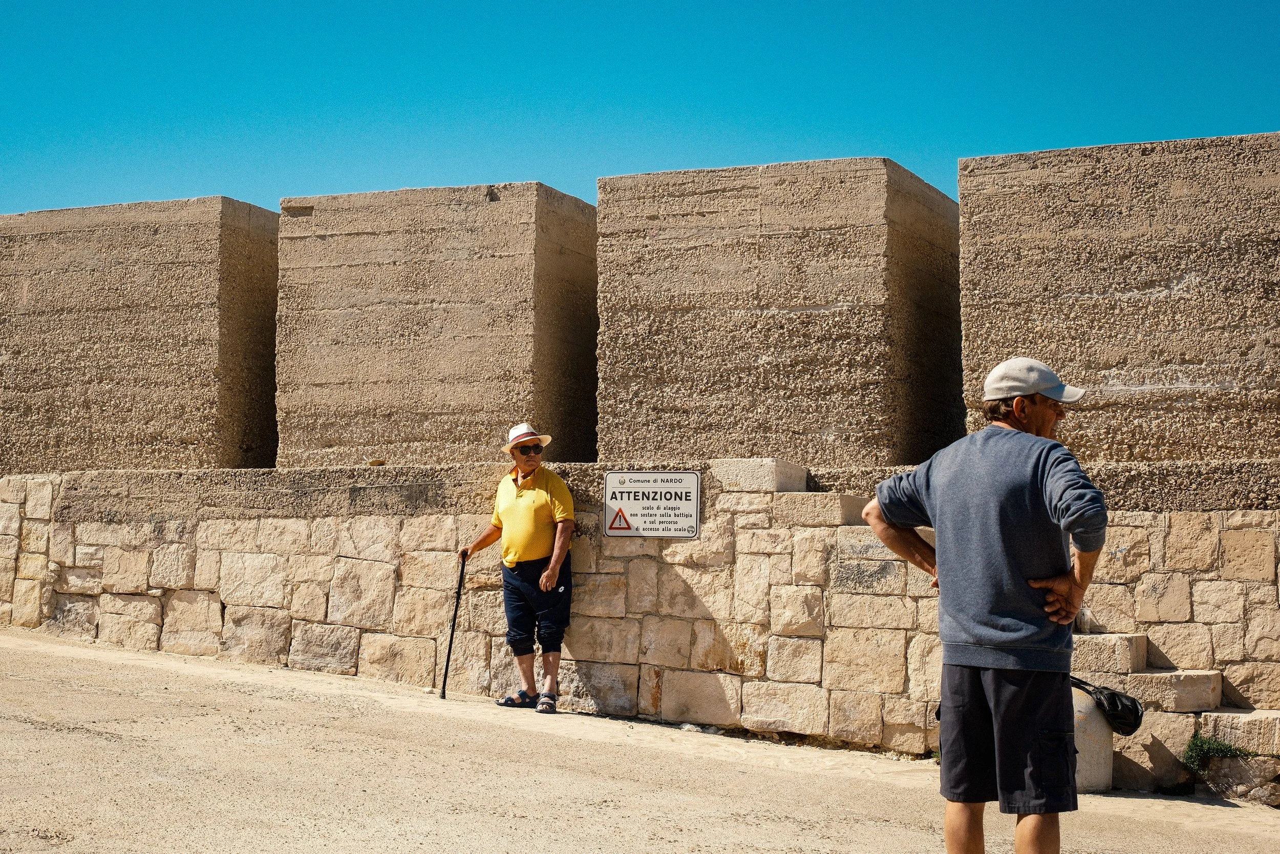 Tourists exploring ancient stone ruins under a clear blue sky, with a sign warning about stairs and pathways.