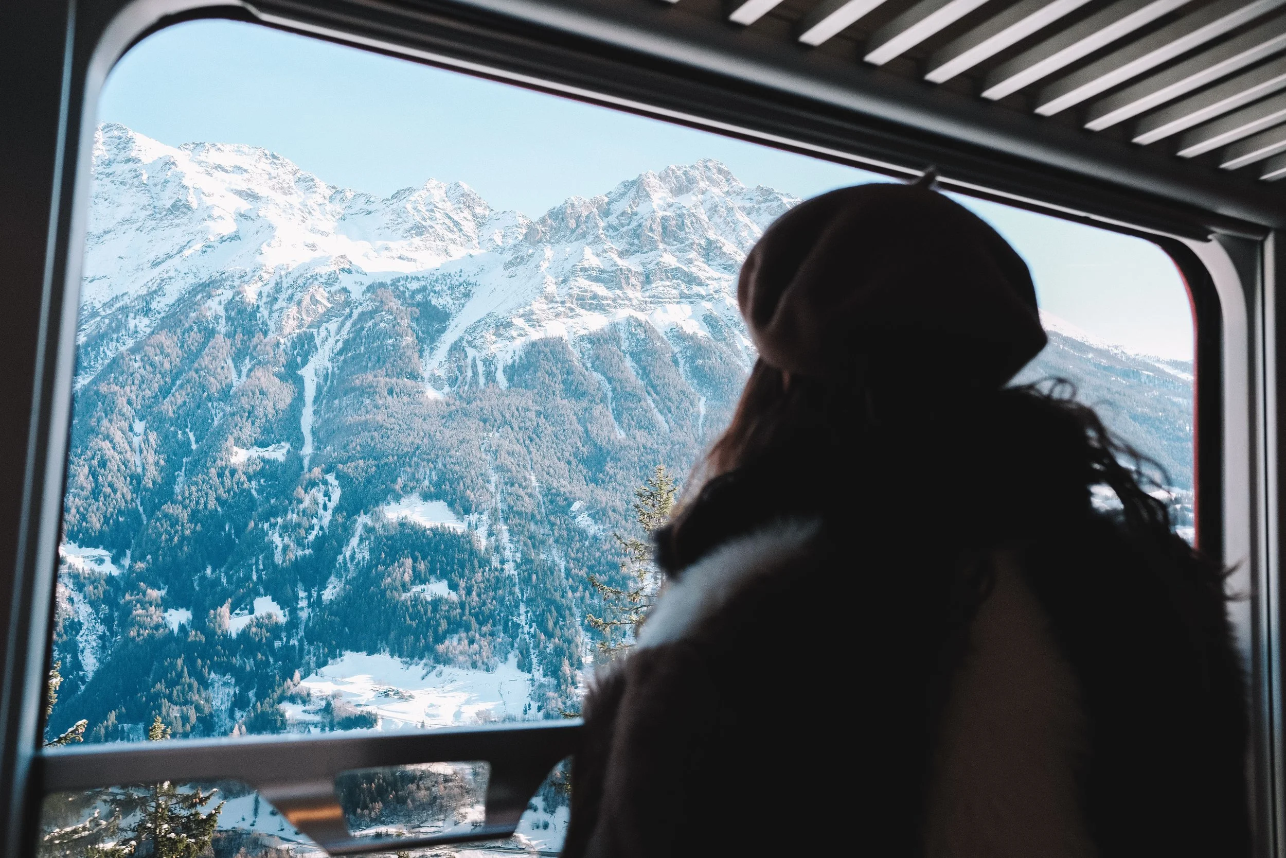 A person in a fur-lined coat and beret, looking out a train window at snowy mountains and forested landscape.