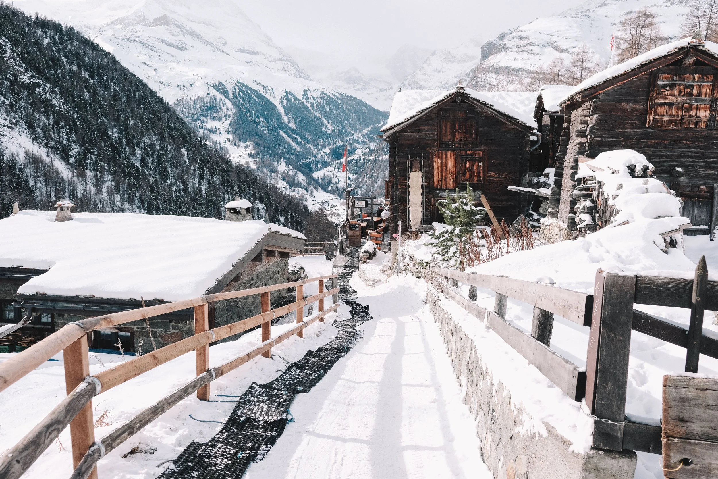 Snow-covered alpine village with wooden cabins and mountains in the background.
