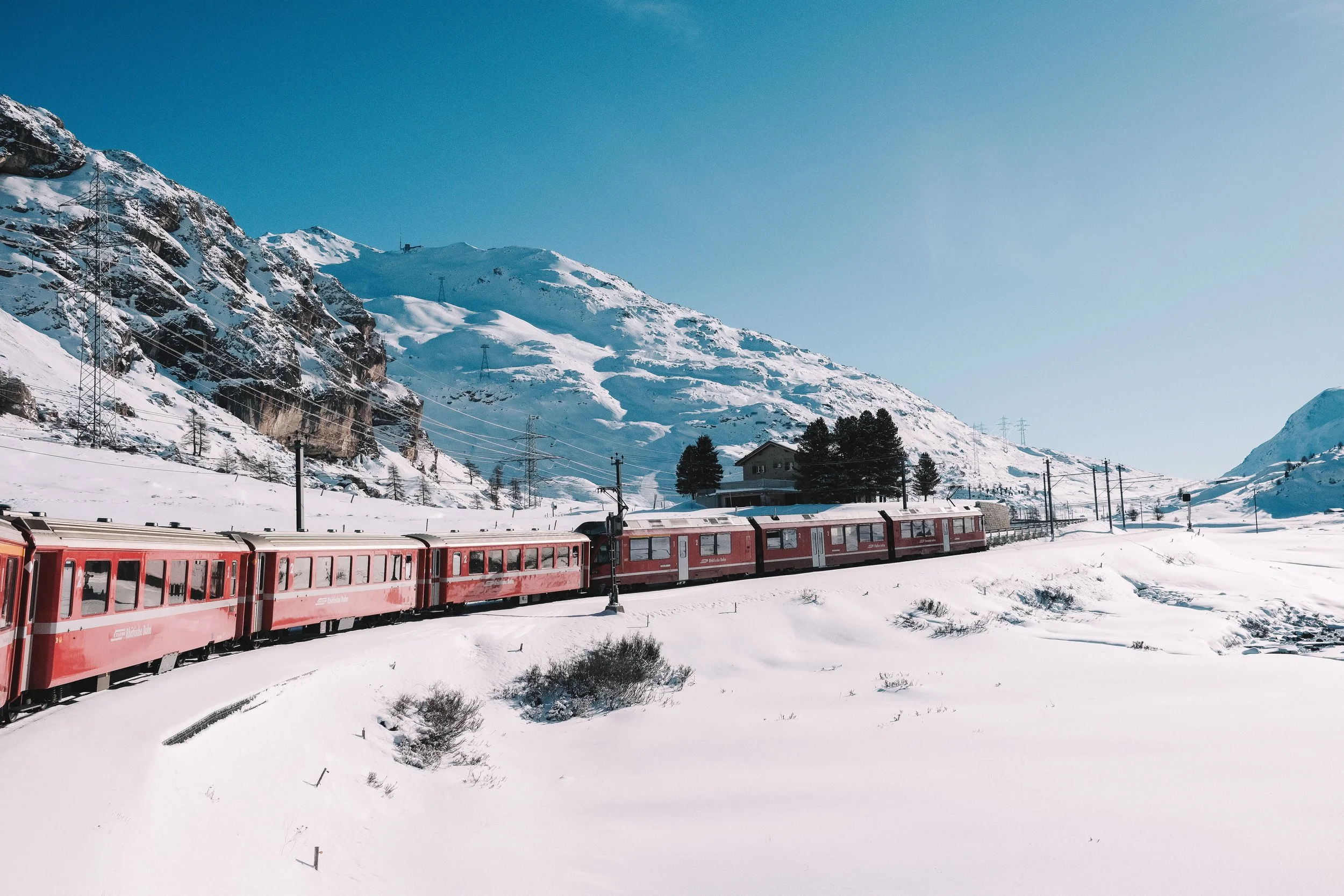Red train traveling through snowy mountainous landscape under clear blue sky.