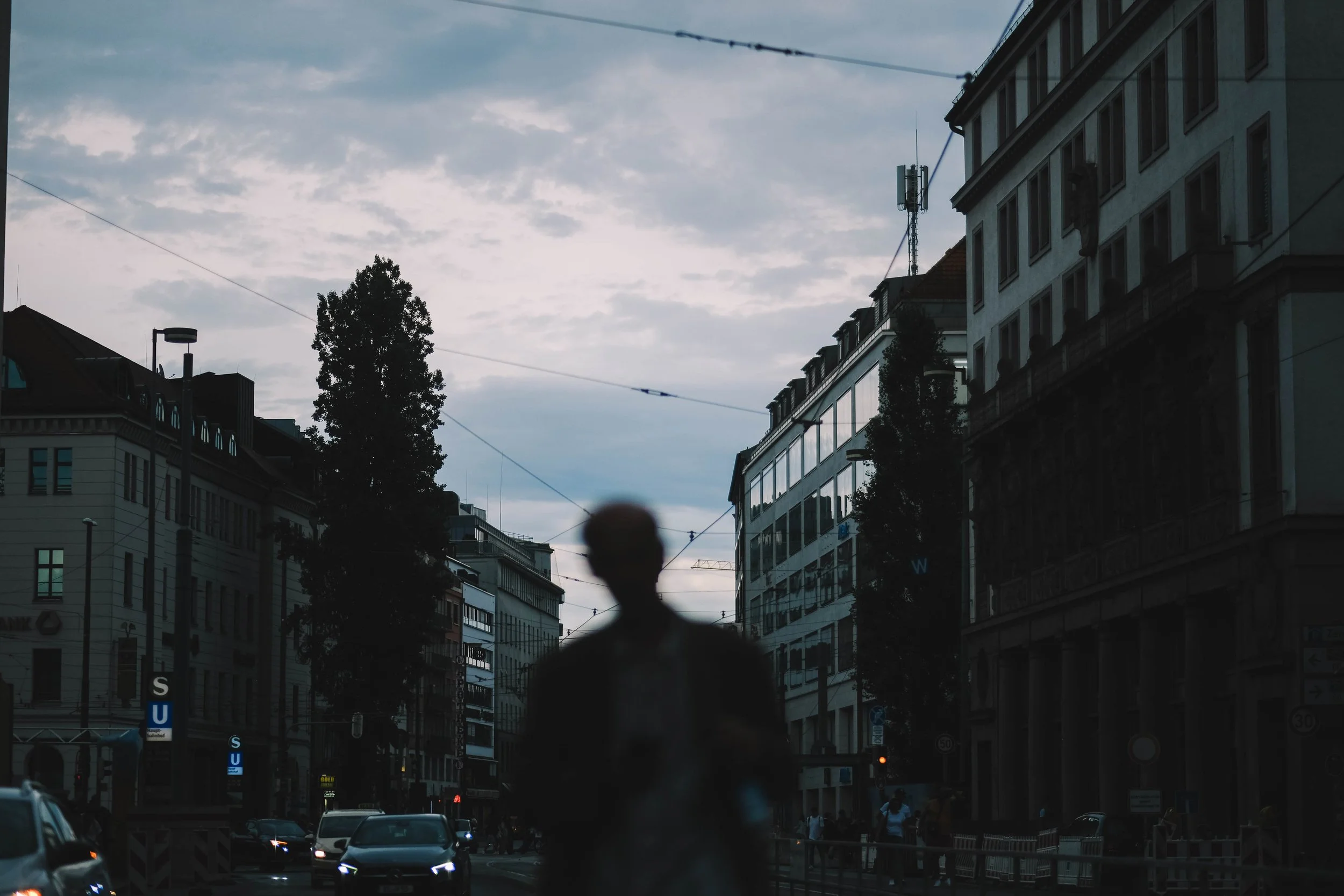 Blurred silhouette of a person walking on a city street at dusk, with cars and buildings visible in the background under a cloudy sky.