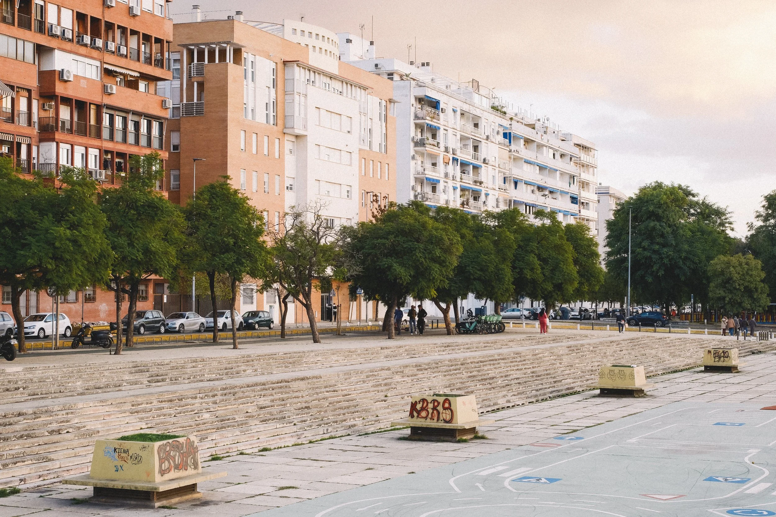Urban park with a basketball court, concrete benches with graffiti, steps, trees, parked cars, and multi-story residential buildings in the background, during daylight.
