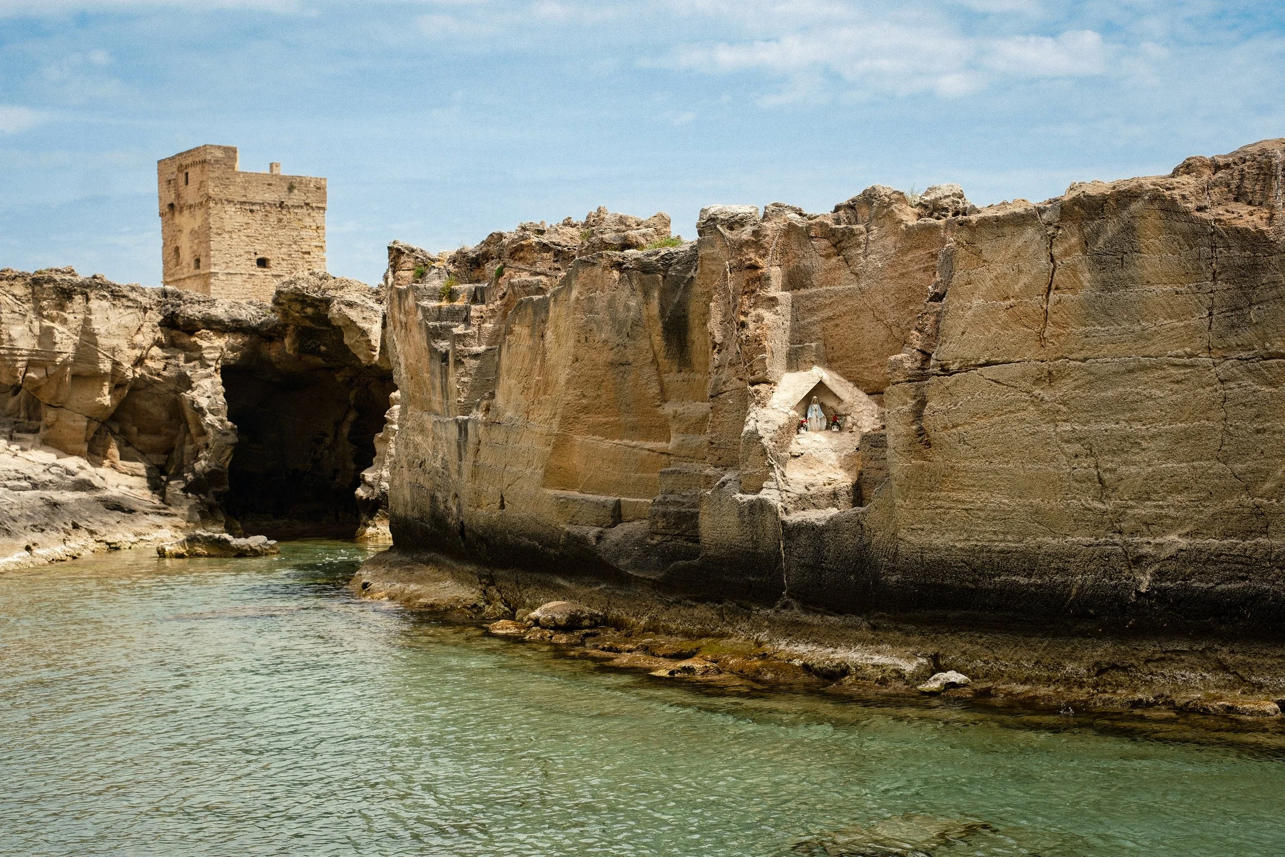 Ancient stone fortress atop a rocky cliff beside a river, with a small shrine containing religious figurines at the base of the cliff, under a blue sky with scattered clouds.
