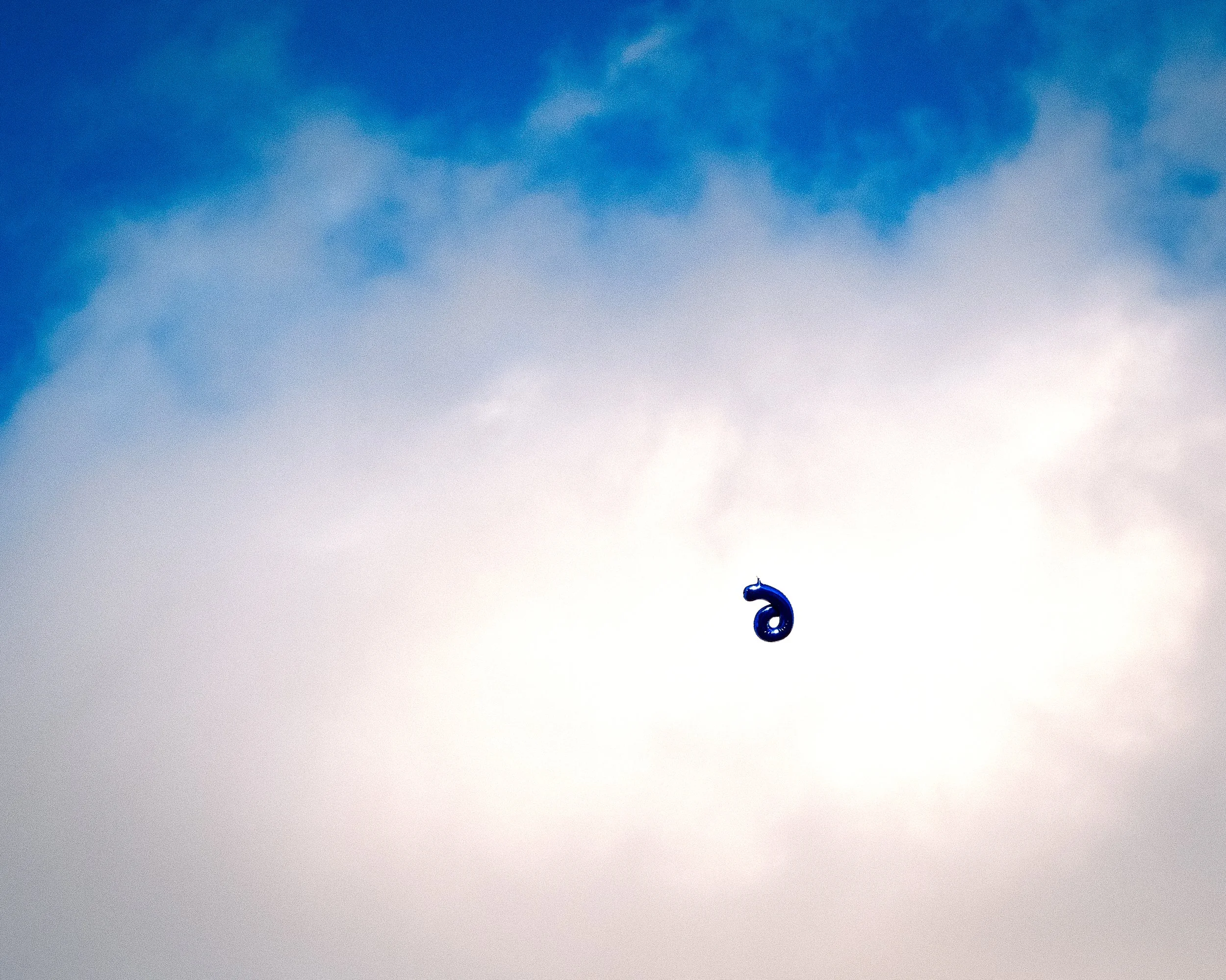 A blue number 3 balloon floating in the sky amid clouds with areas of blue sky visible.